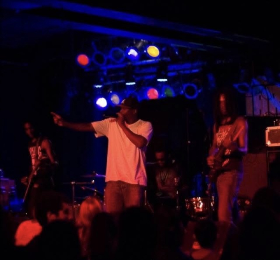 Performer with microphone on stage, band members playing instruments, colorful stage lights, audience in foreground.
