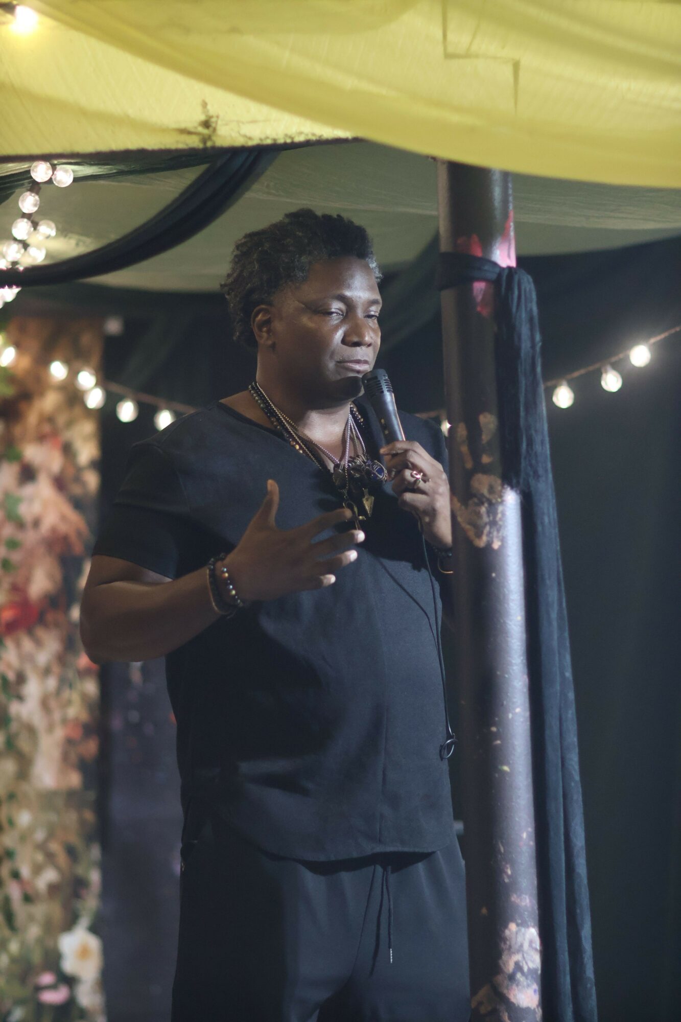 Person with dark hair and dark clothing speaking into a microphone at an event, with string lights and a yellow canopy in the background.