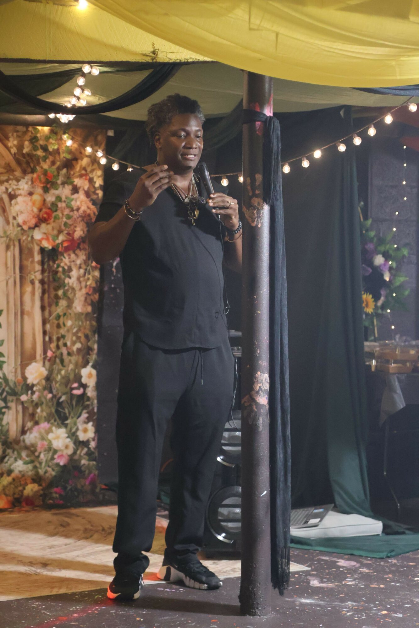 Person in dark suit speaking into a microphone on stage with string lights and floral backdrop.