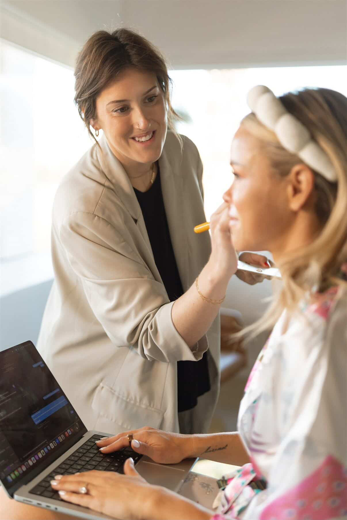 Woman feeding a young girl with a spoon, girl wearing a headband, in a bright room, laptop on table, smiling.