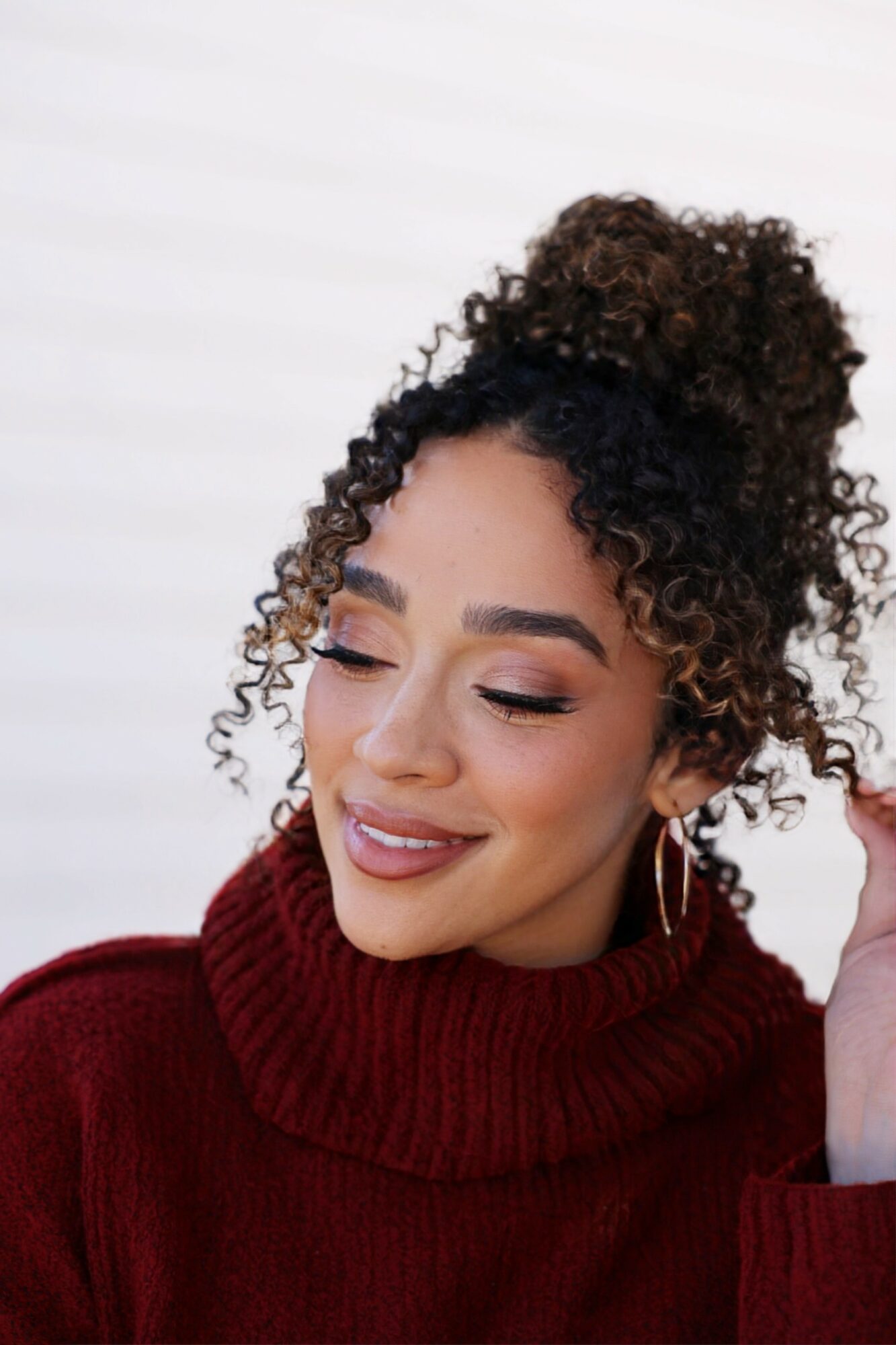 Smiling woman with curly hair in a high bun, wearing a red turtleneck, against a light background.