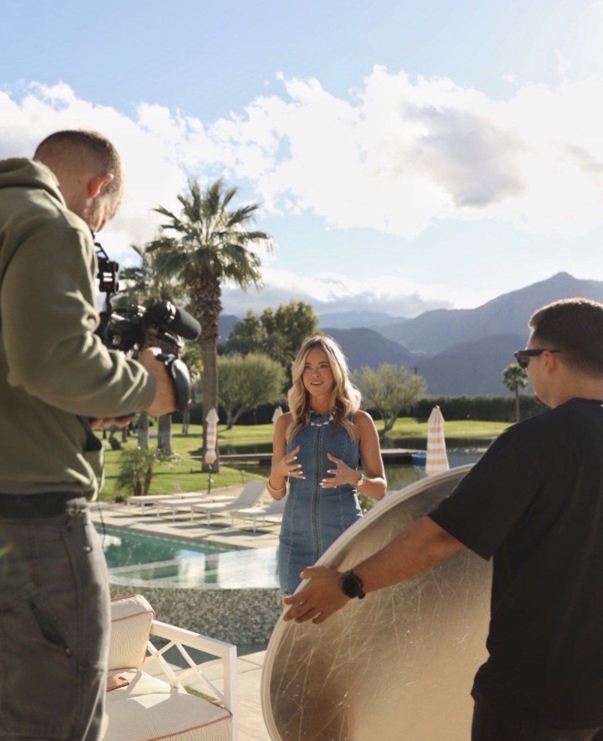 A woman is being filmed outdoors with mountains, palm trees, and a pool in the background, during daytime.