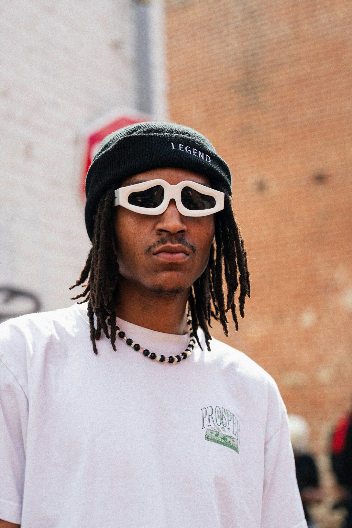 Young person with dreadlocks wearing a black beanie, white sunglasses, a white shirt, and a beaded necklace outdoors.