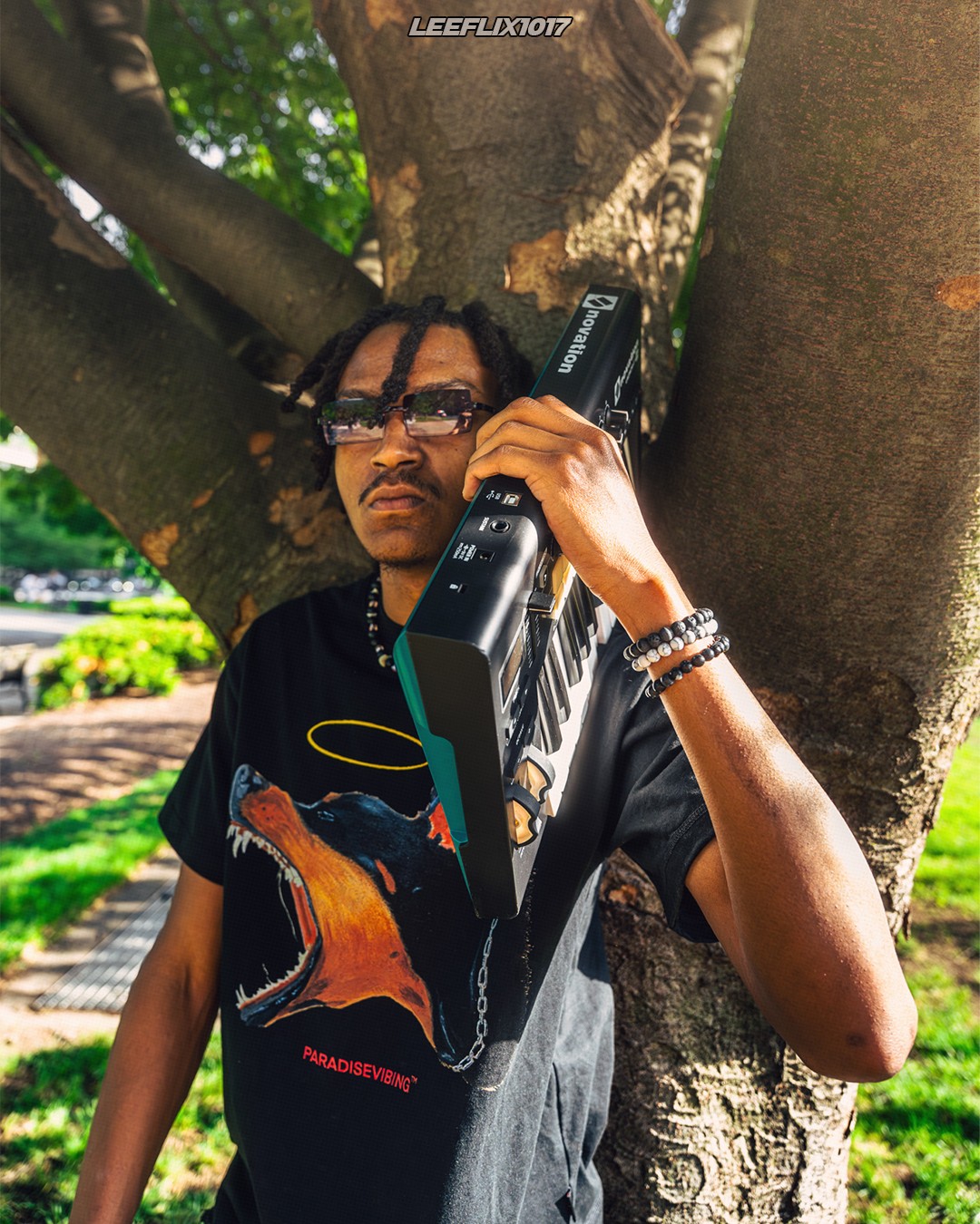 Person with sunglasses and bracelets holding a guitar case, standing against a tree outdoors.