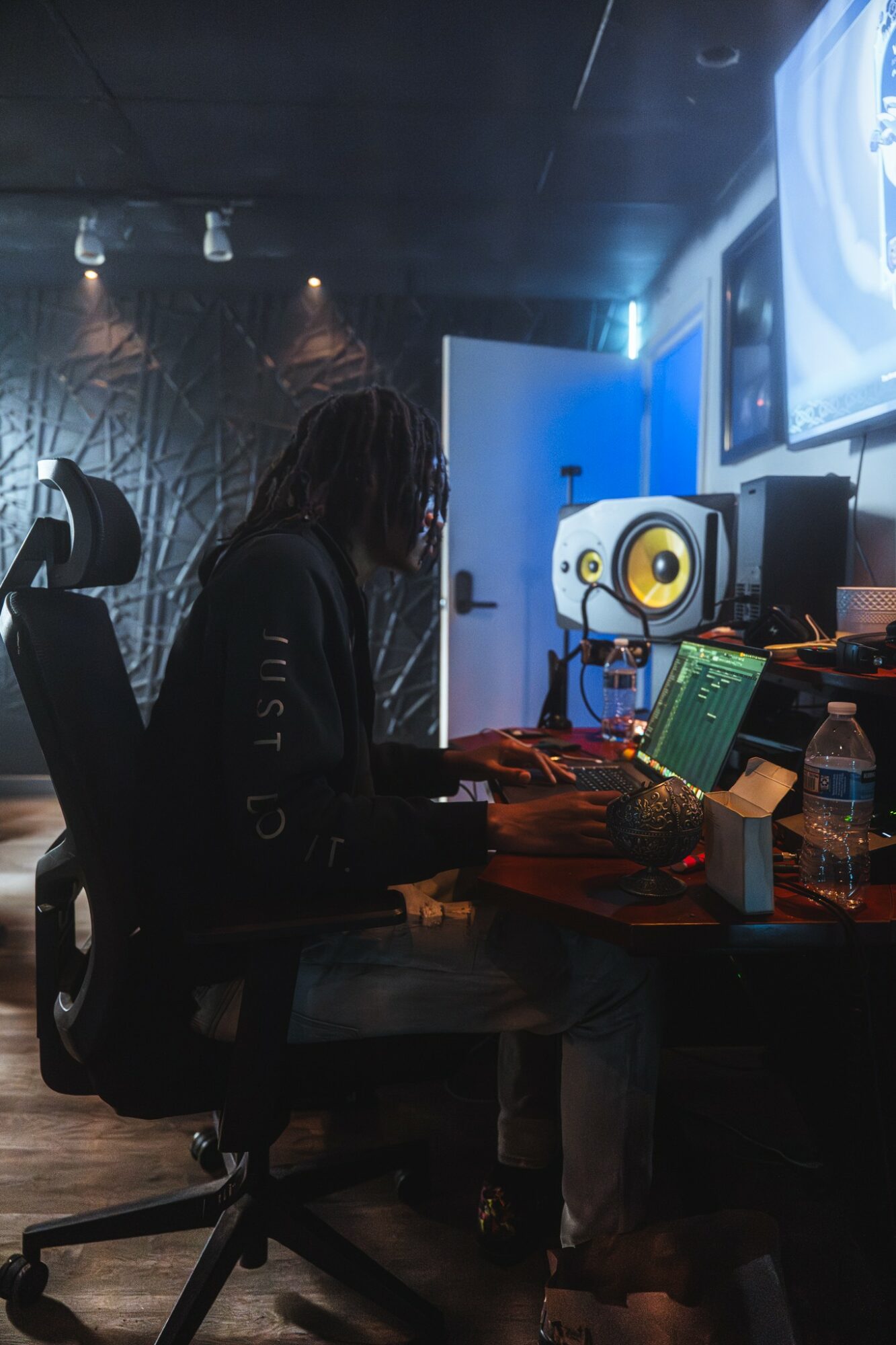 Person working at a desk with audio equipment and a computer in a dark room, illuminated by screens and ambient lighting.