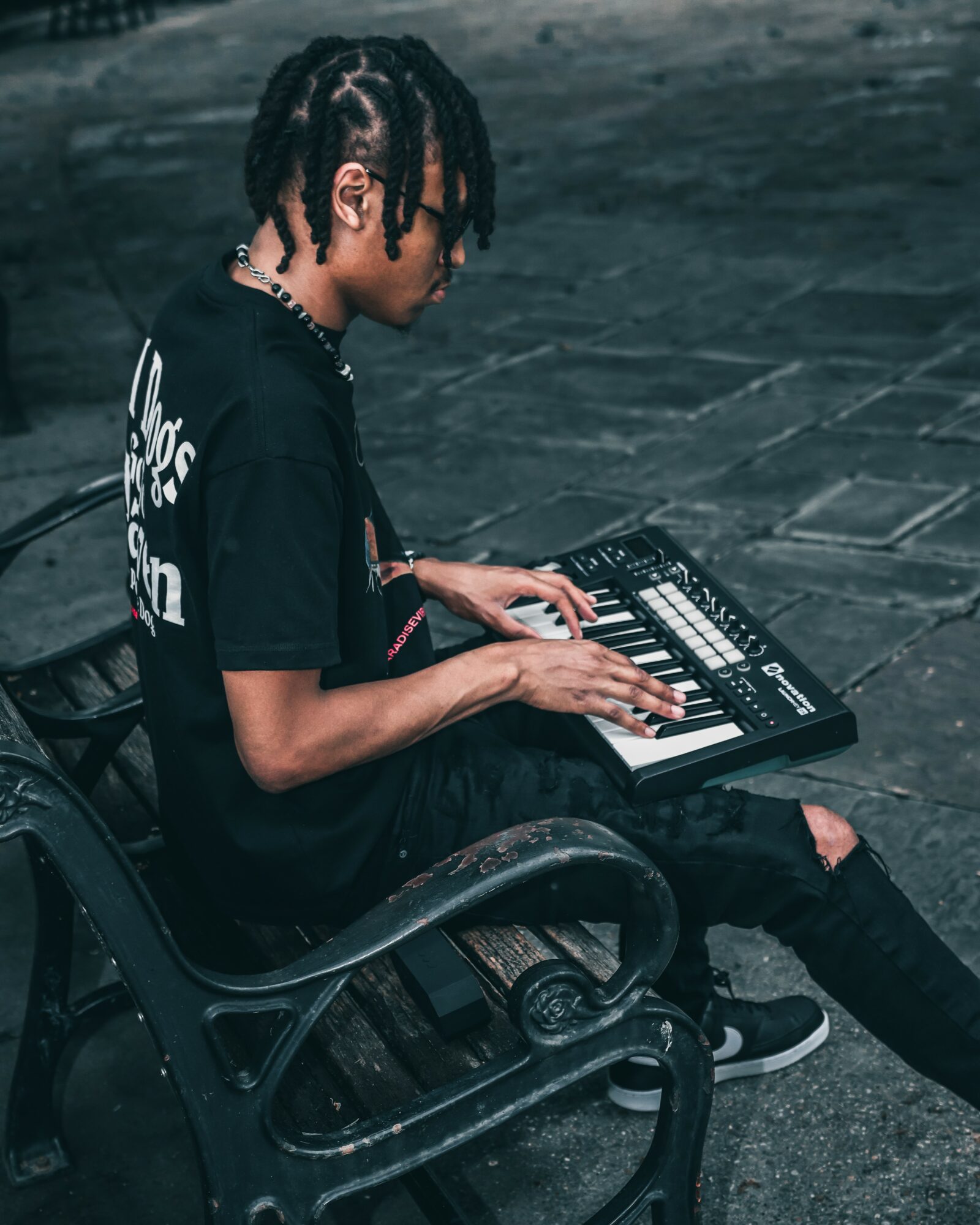 Person with dreadlocks playing keyboard on a bench outdoors, wearing a black T-shirt and ripped jeans.