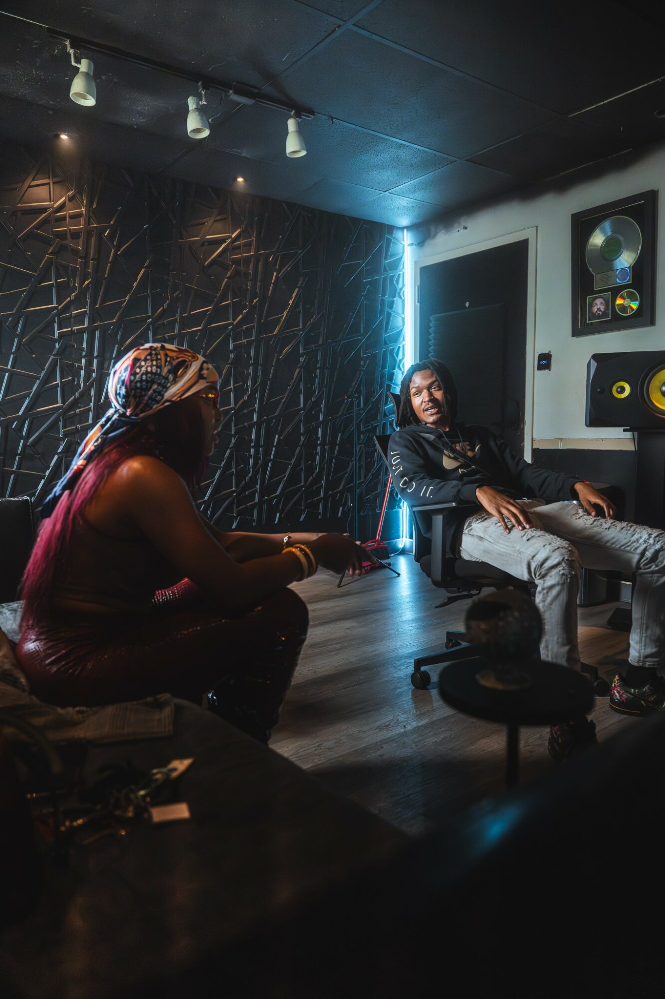 Two women sit in a dimly lit room with music awards on the wall, one with a headscarf and the other with long hair.
