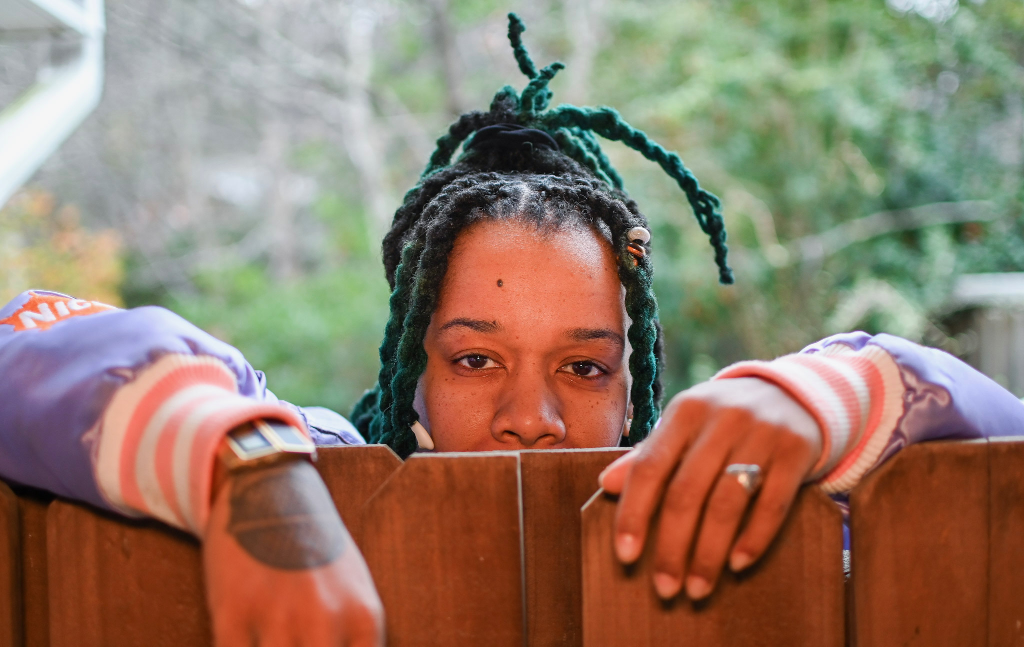 Person with braided hair resting their head on a wooden surface, looking directly at the camera, outdoors with greenery in background.