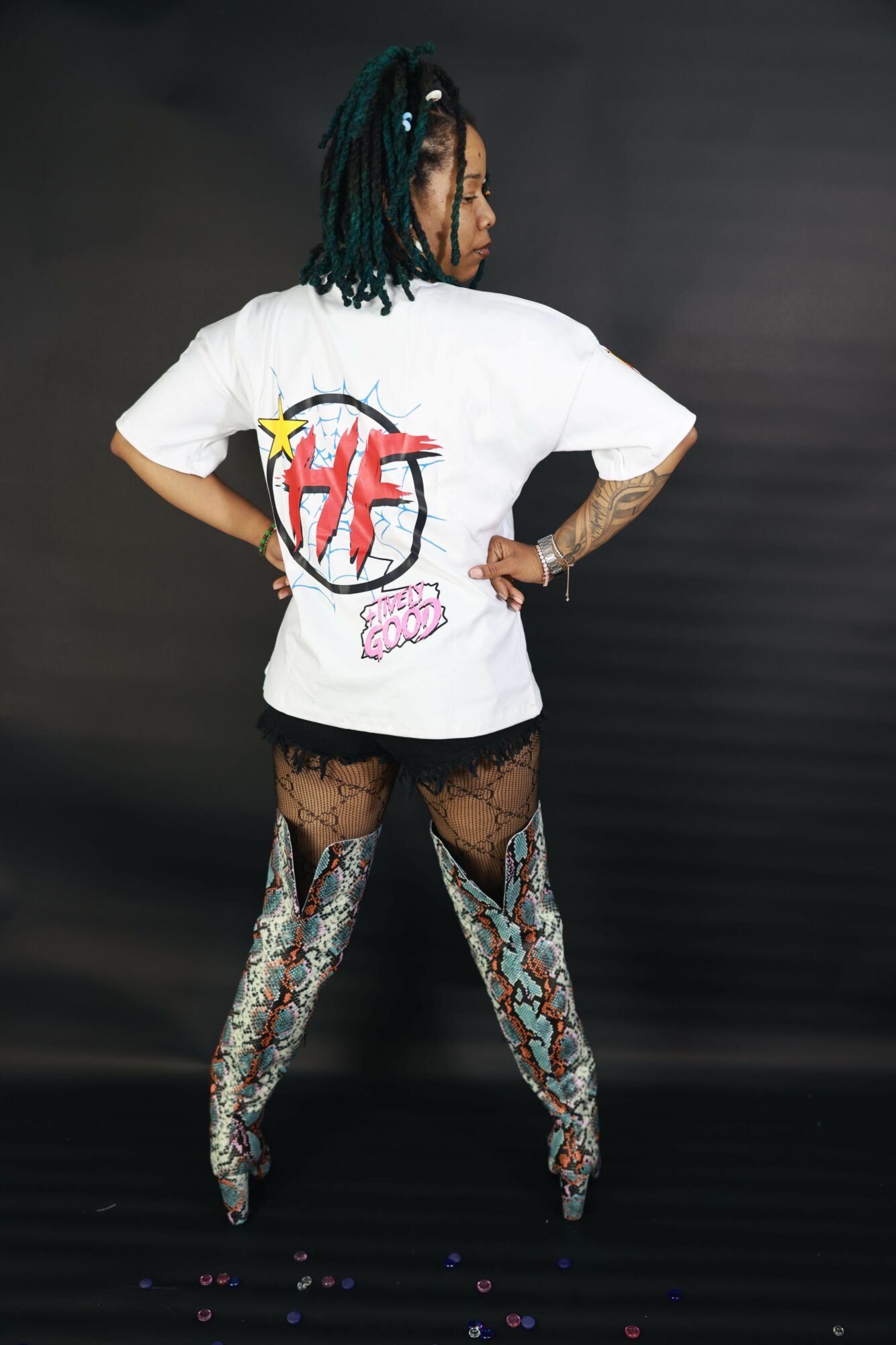 Person with long dreadlocks wearing a white T-shirt with a red and pink graphic, patterned thigh-high boots, and black shorts, standing sideways against a dark background.