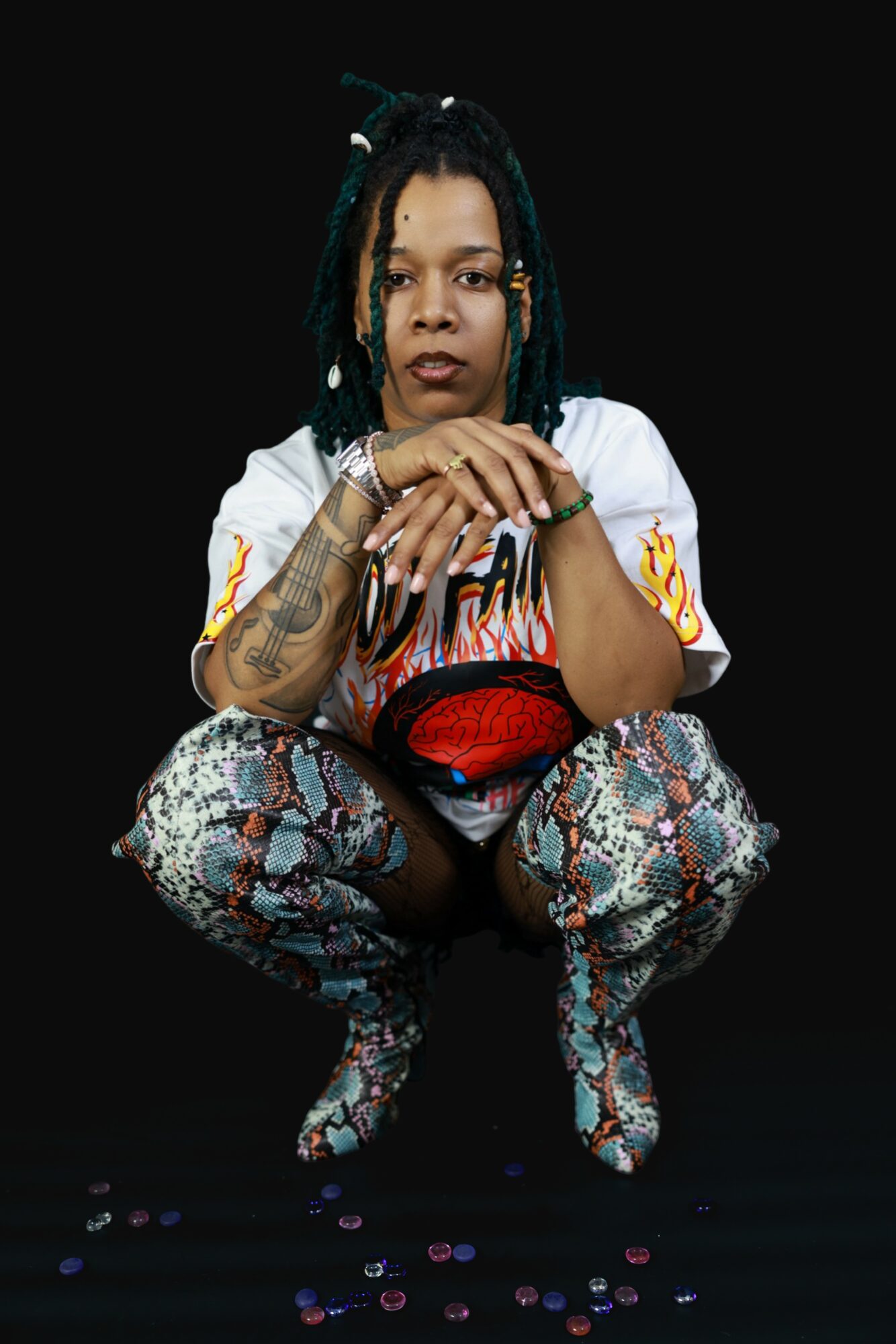 Person with dreadlocks crouching, wearing patterned pants and a white shirt, against a black background.