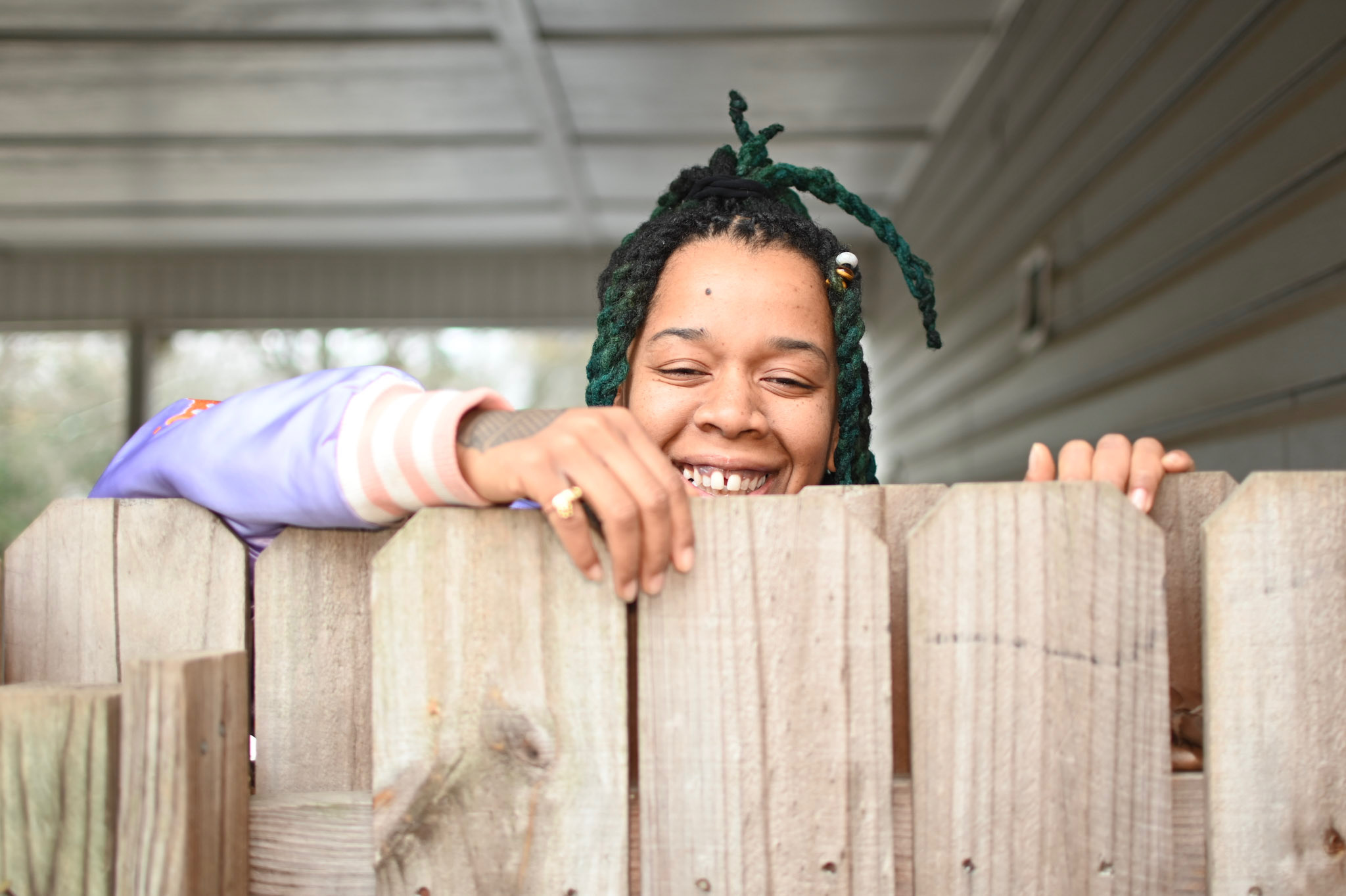 Person with green dreadlocks smiling and peeking over a wooden fence outdoors.