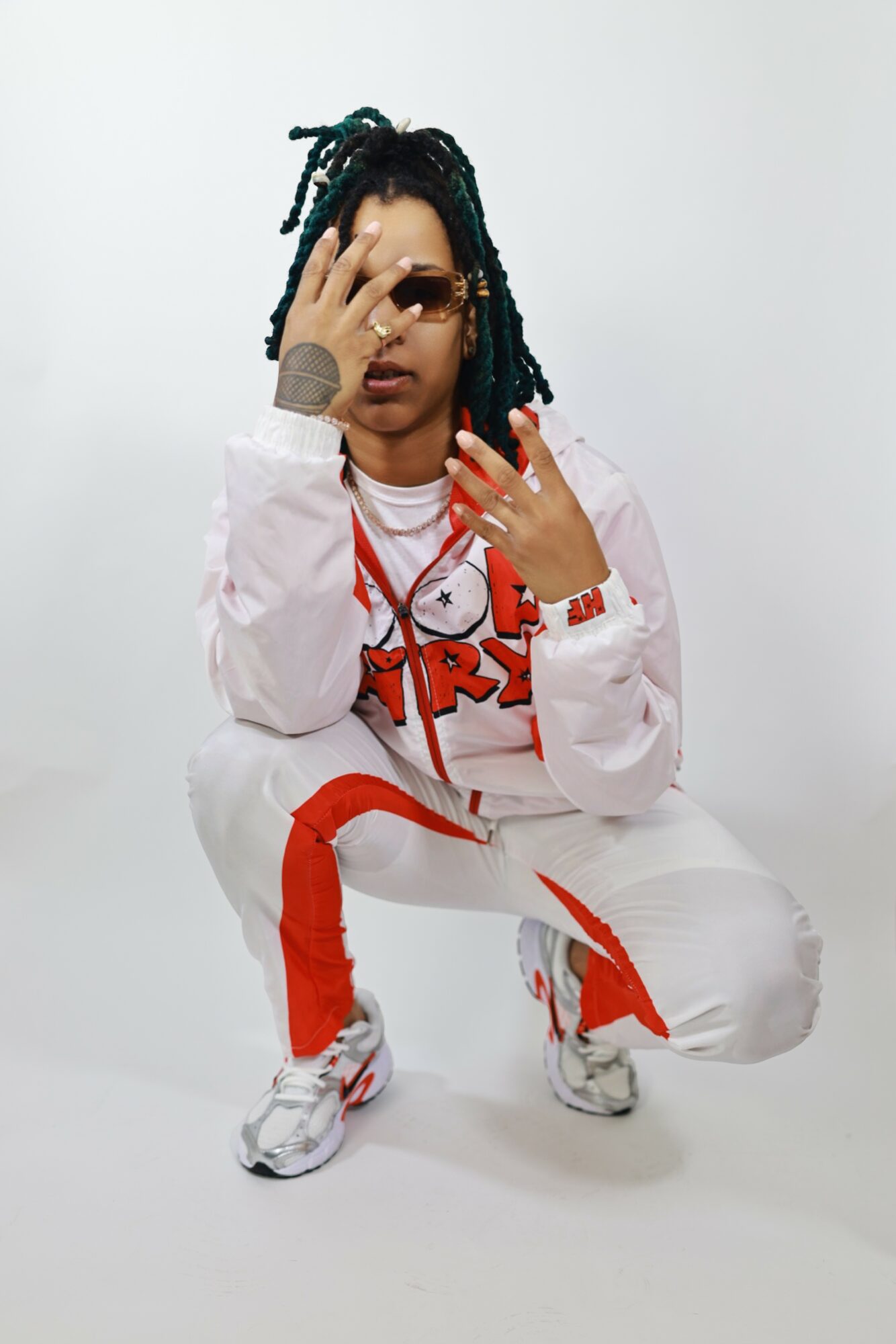 Person with dreadlocks crouching, wearing white and red clothing, sunglasses, and jewelry, against a plain white background.