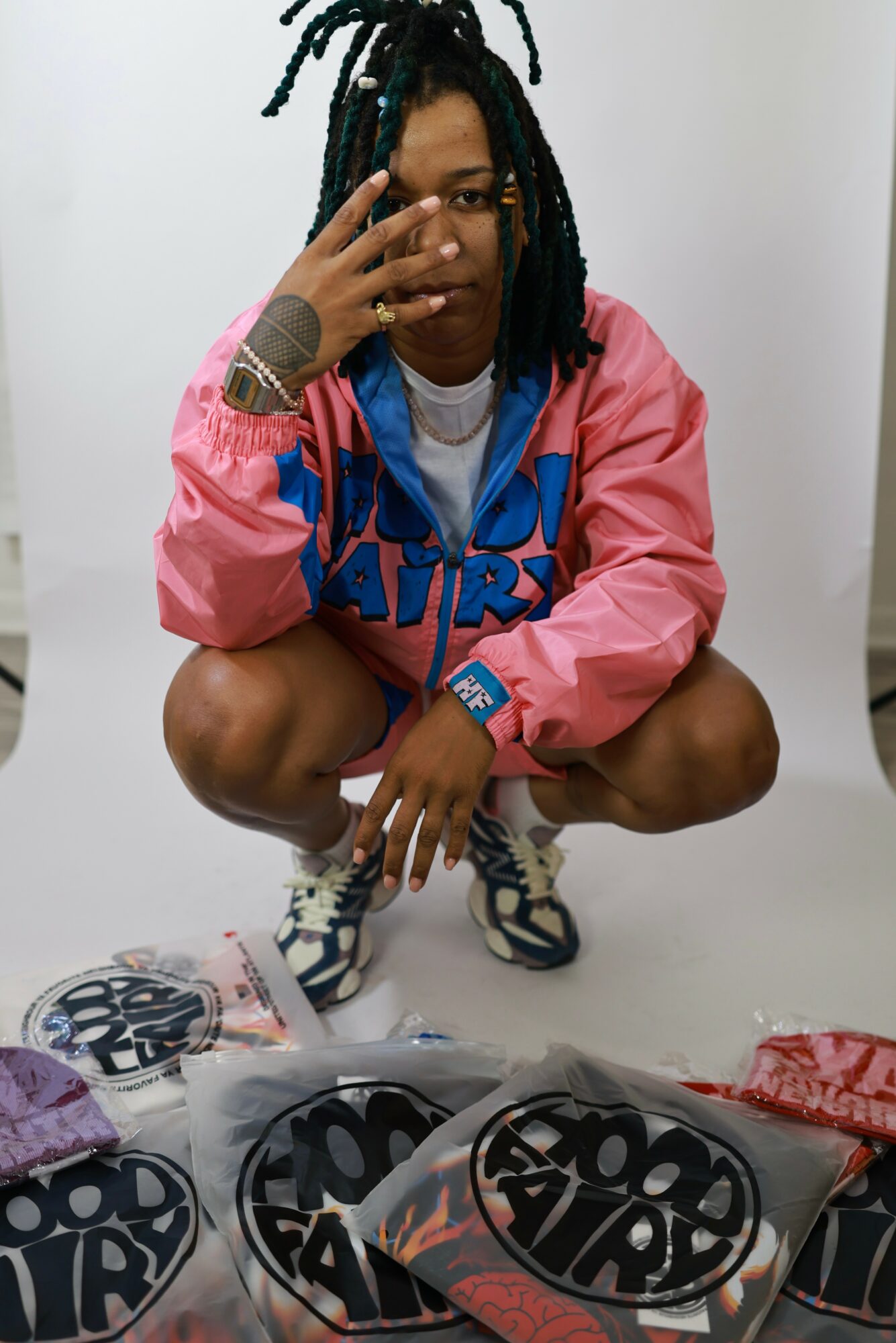 Person crouching with dreadlocks, wearing a pink and blue jacket, white shirt, and sneakers, in front of a white background.