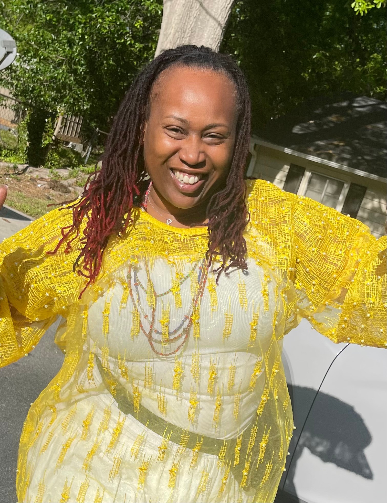 Smiling woman with dreadlocks wearing a yellow dress outdoors, trees and a house in background.