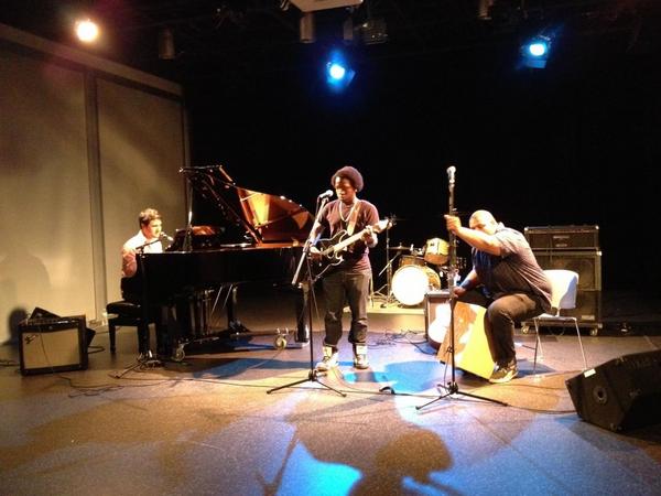 Three musicians perform on stage with a piano, guitar, and drums, under stage lights, in a dark setting.