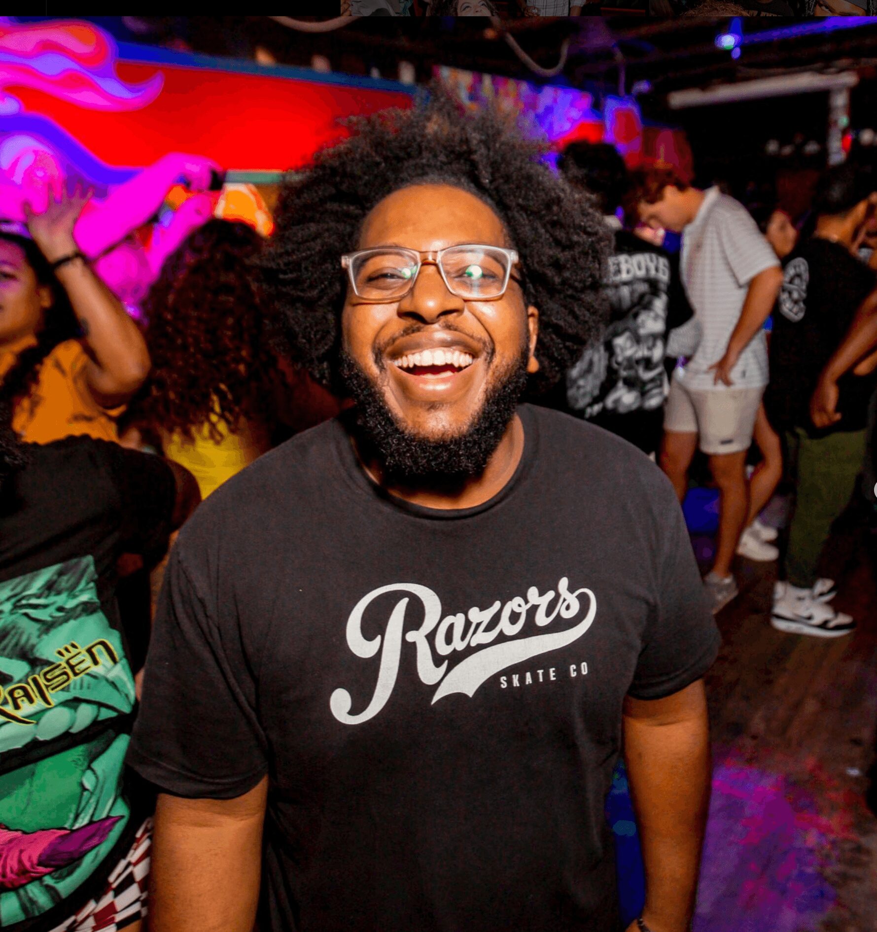 Man with glasses and curly hair smiling at a lively party or club scene.