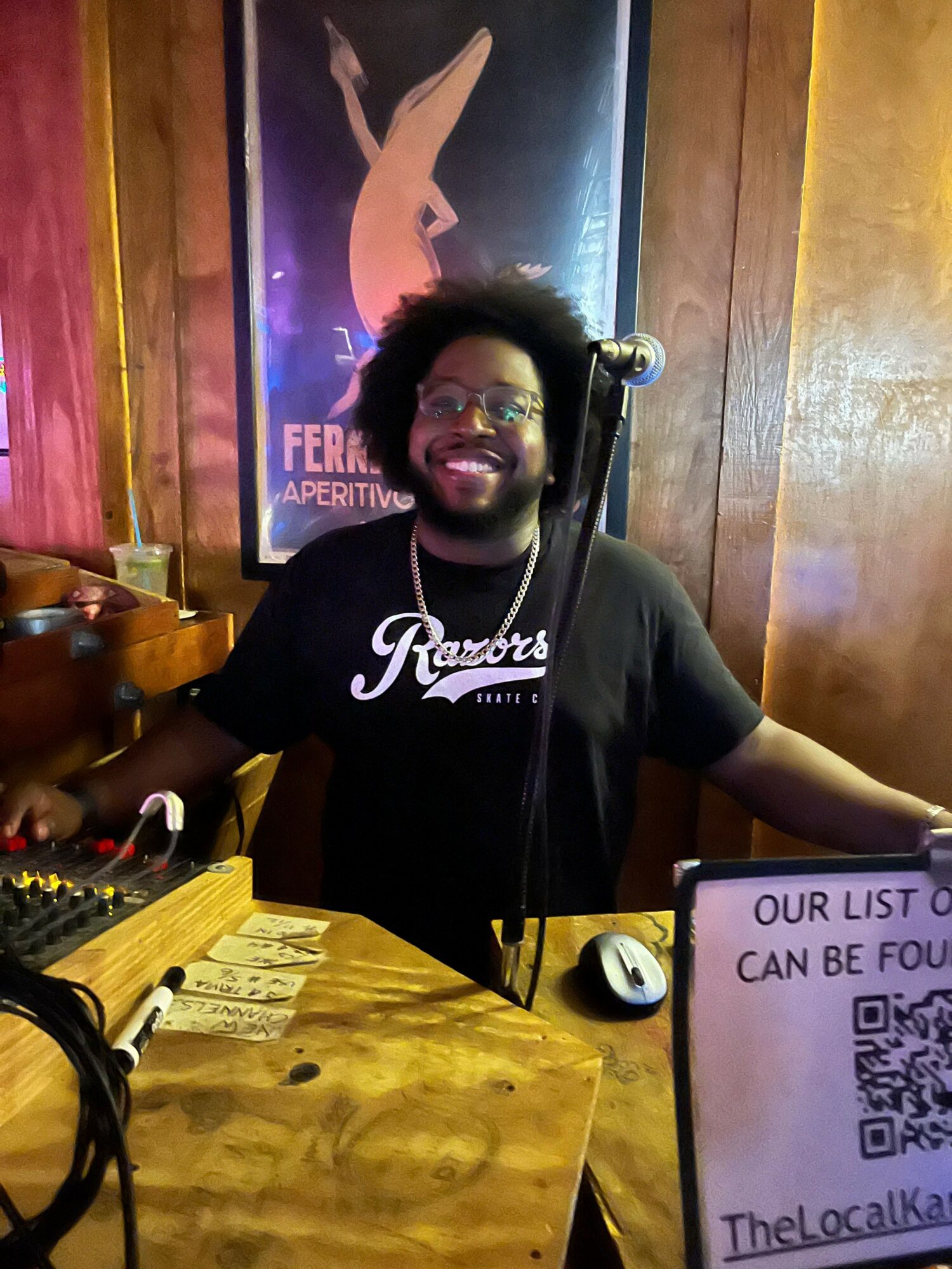 Person with glasses and beard smiling, sitting at a wooden table with a pink menu, in a dimly lit setting.