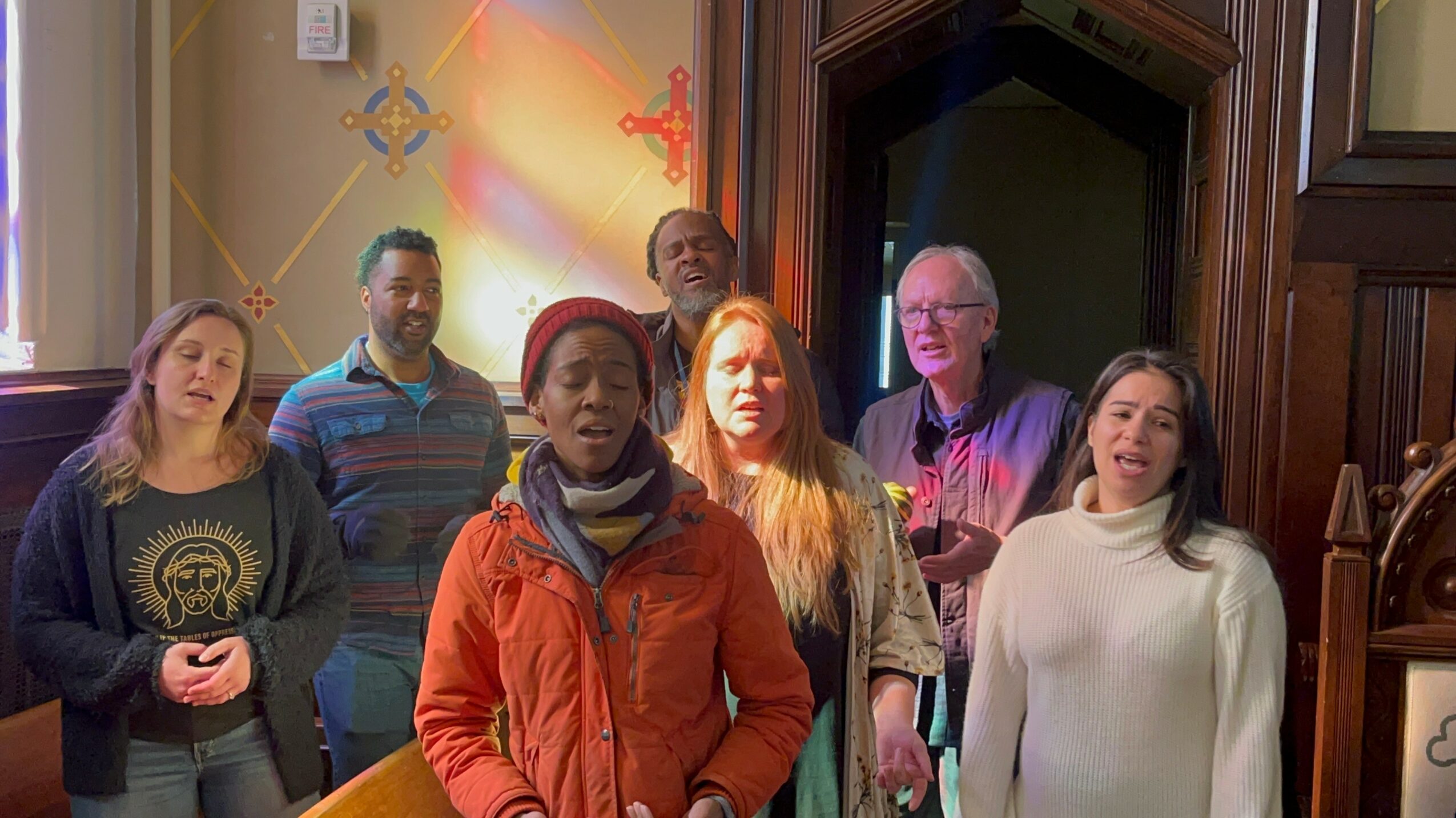 Group of seven people standing indoors, some looking at the camera, with colorful wall and wooden panel background.