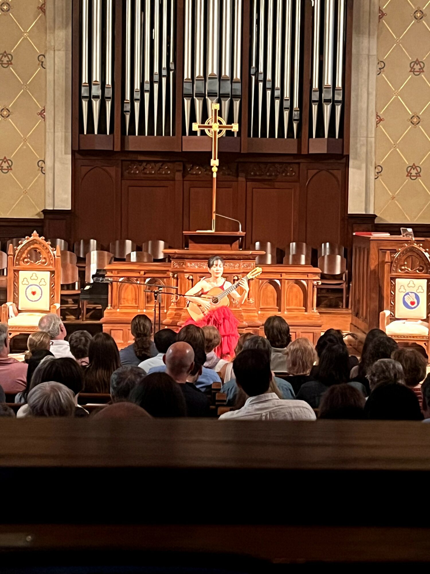Person playing guitar on stage in front of an audience, with a large pipe organ and decorative wall behind.