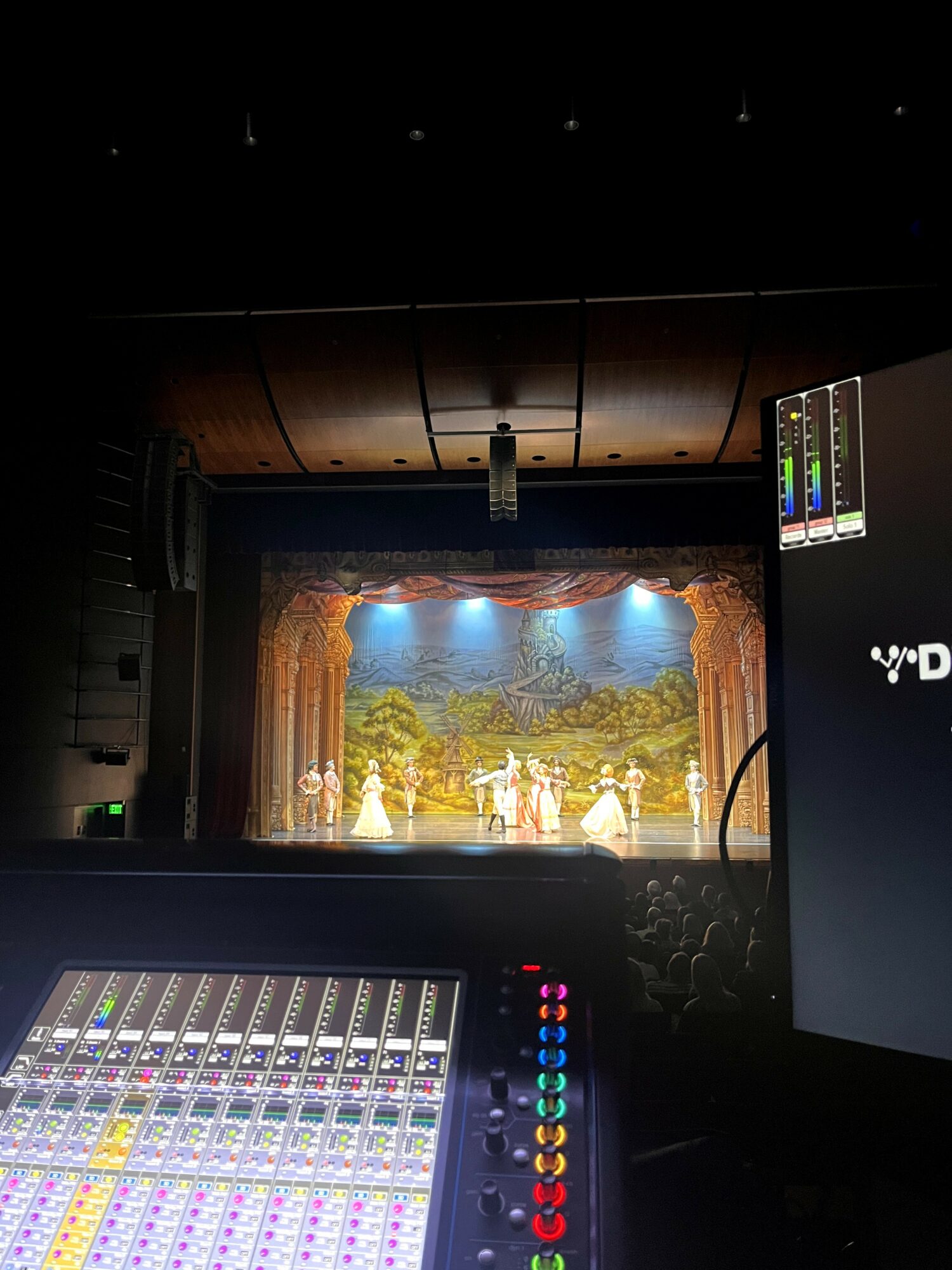 The stage shows performers in costumes with a scenic backdrop, viewed from the control room with audio equipment visible.
