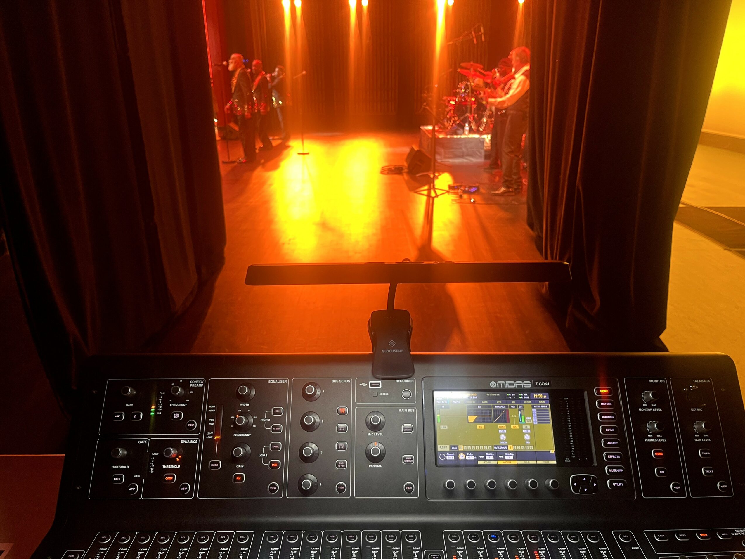 Stage with performers illuminated by warm orange lighting, viewed from behind a sound mixing console in the foreground.