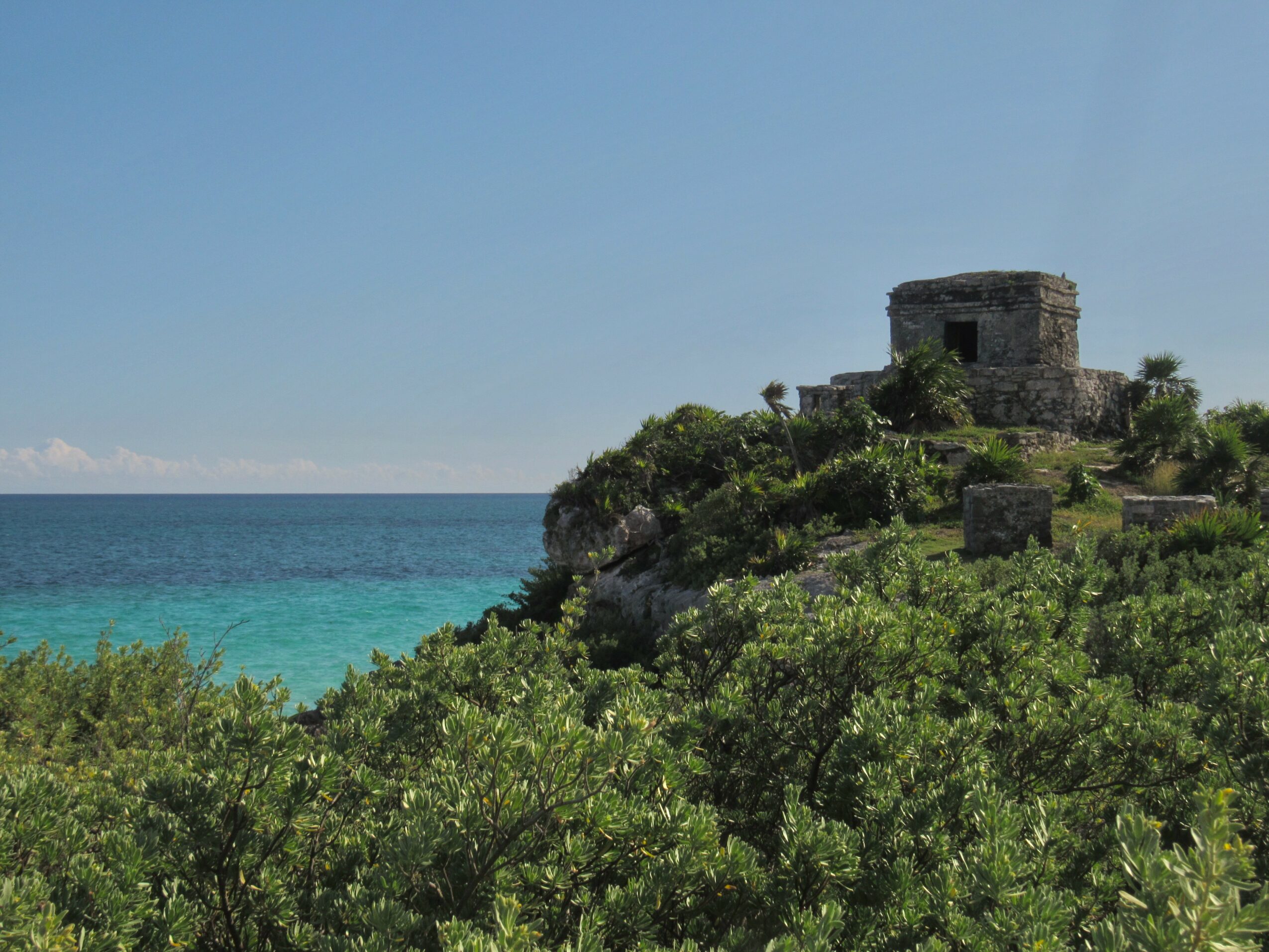 Stone building on a hill overlooking the ocean with green bushes in foreground, clear blue sky above.