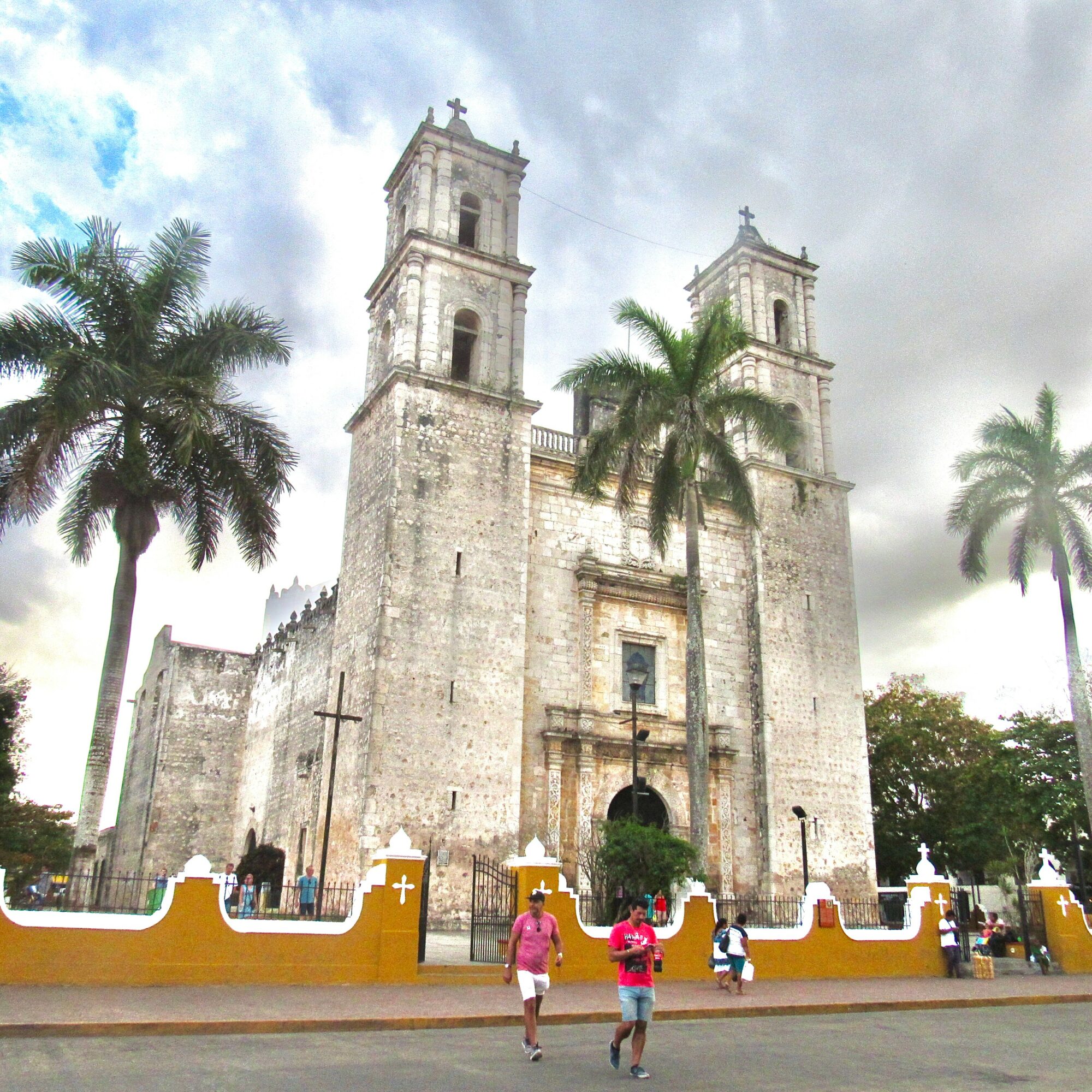Historic church with two towers, surrounded by palm trees and a fence, under cloudy sky, with people walking nearby.