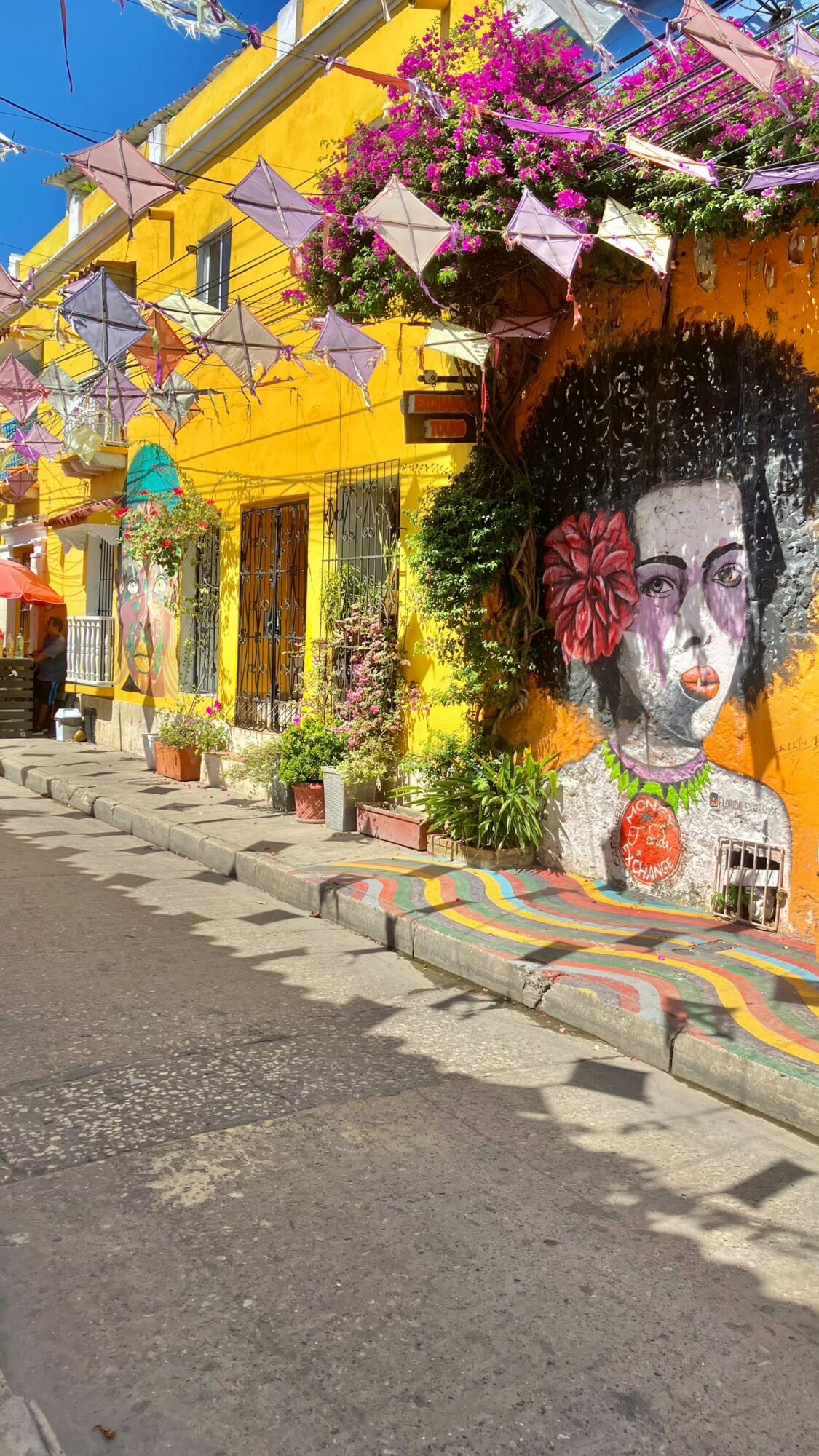 Street scene with colorful buildings, murals, and decorations, including a large portrait of a woman with flowers in her hair.