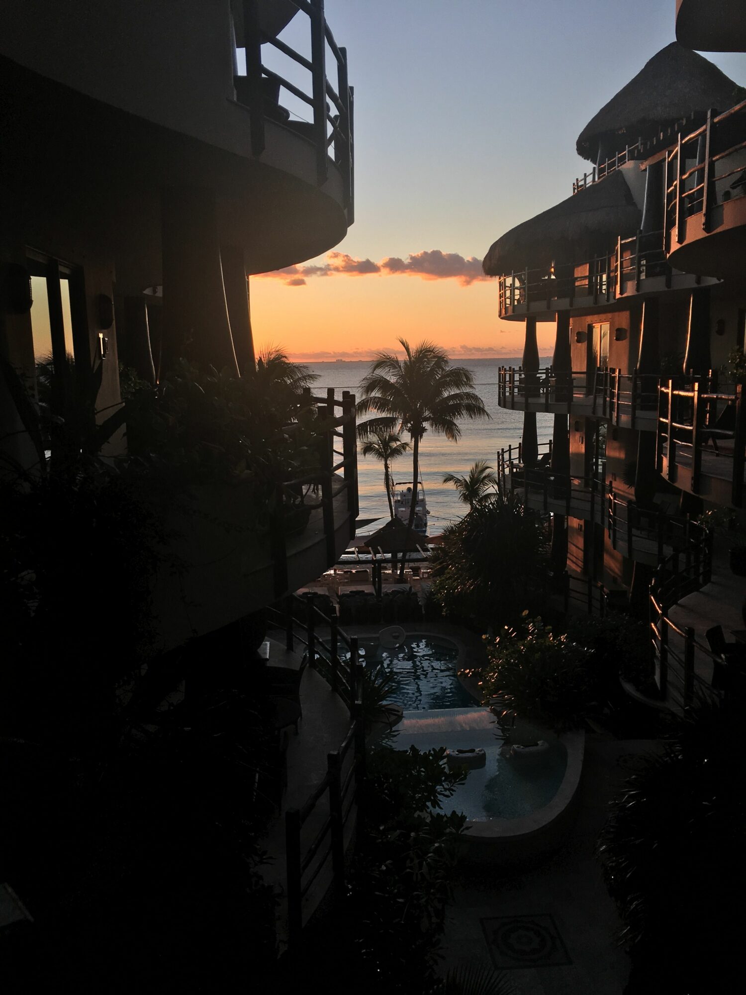 Balcony view overlooking a sunset with palm trees, buildings, and a hot tub, in a tropical setting.