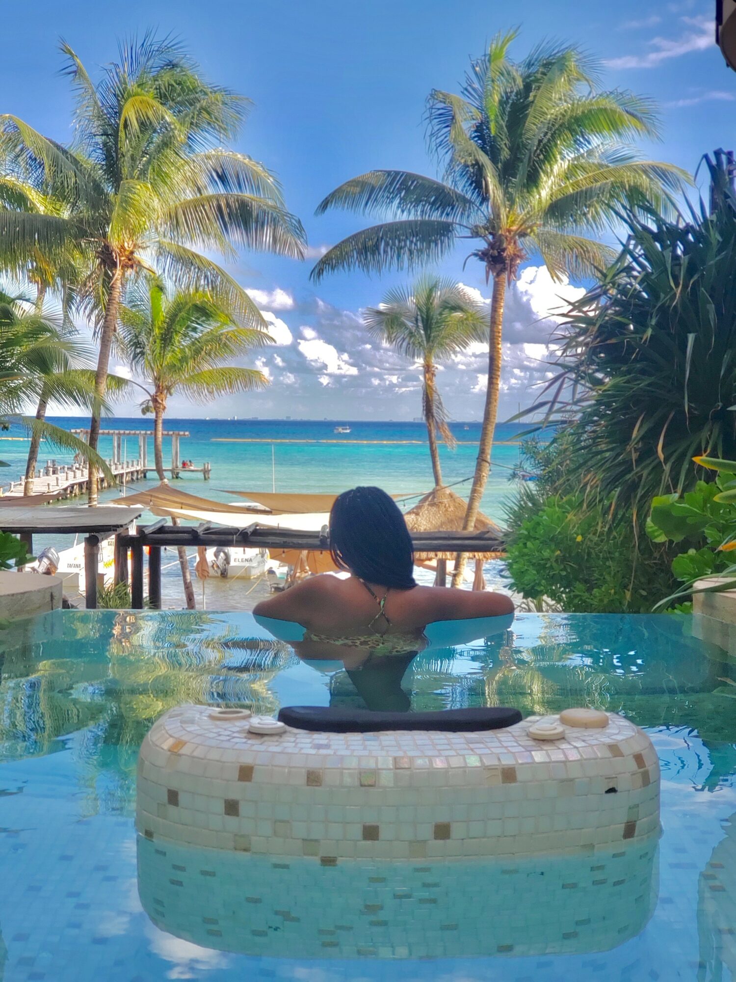 Person relaxing in a pool with palm trees and ocean view in the background, sunny sky, tropical setting.