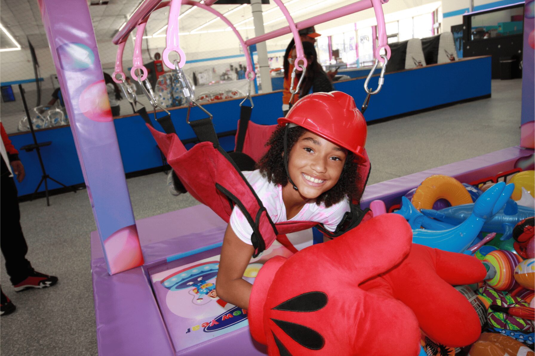 Girl wearing a red helmet and harness, smiling, at an indoor play area with colorful equipment and hanging toys.