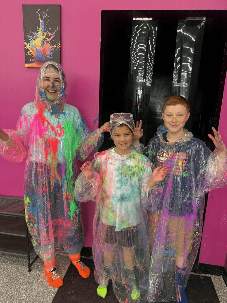 Three people in colorful, paint-splattered protective suits stand in front of a pink wall and black display case, smiling.