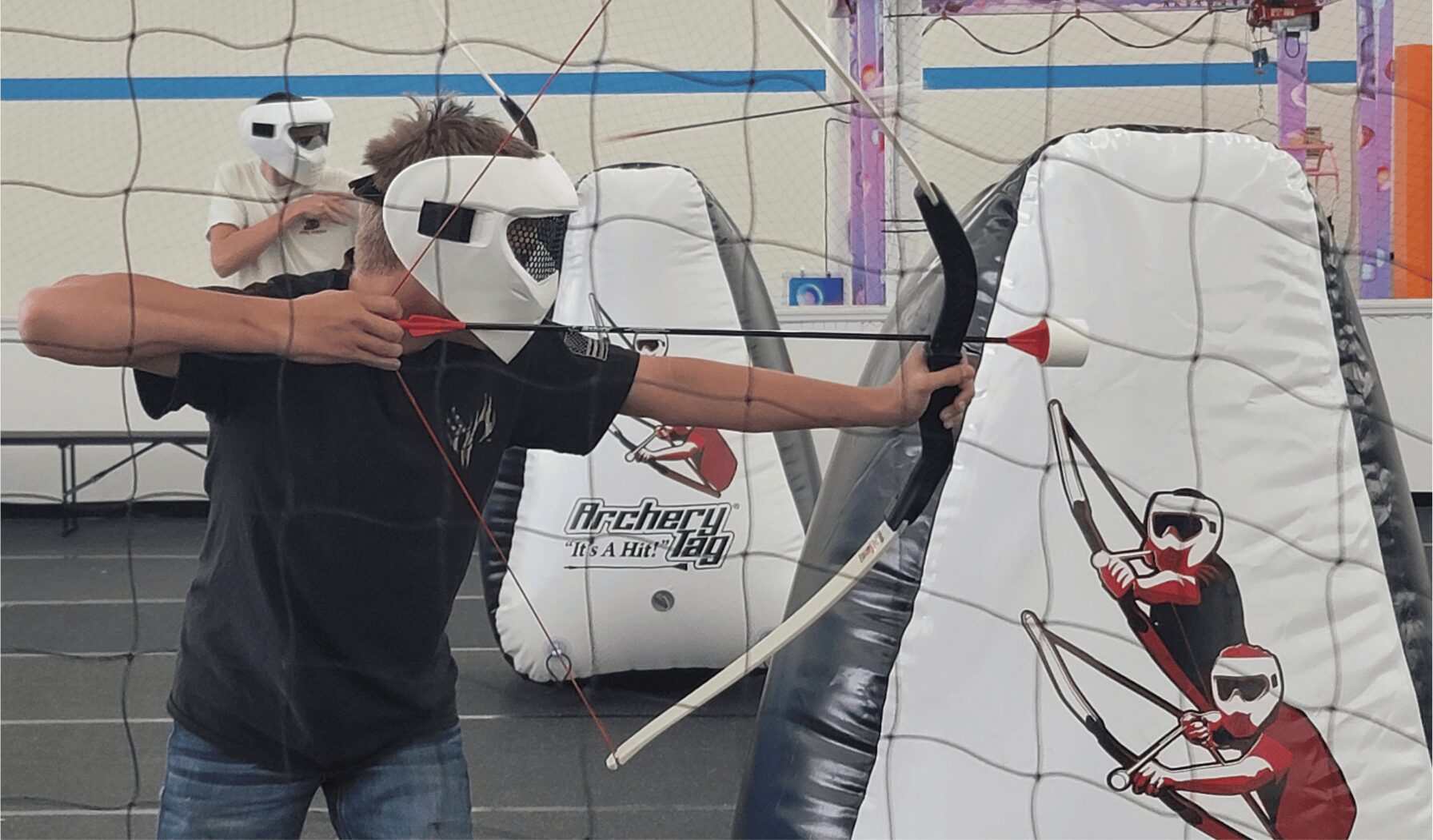 Child wearing a mask and black shirt, holding a bow and arrow at a paintball or archery range, with inflatable targets in background.