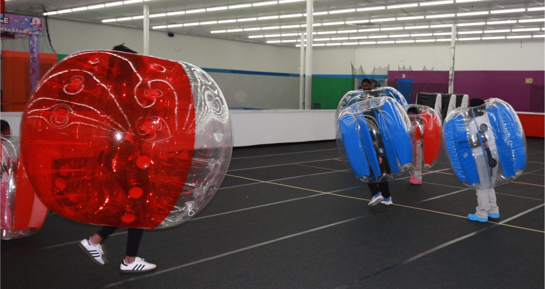People inside large, transparent, inflatable spheres on an indoor sports court, with colorful walls and ceiling lights visible.