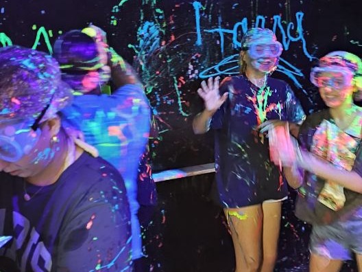 Group of children playing with colorful paint splashes, smiling and waving, in a dark room with bright paint on walls.