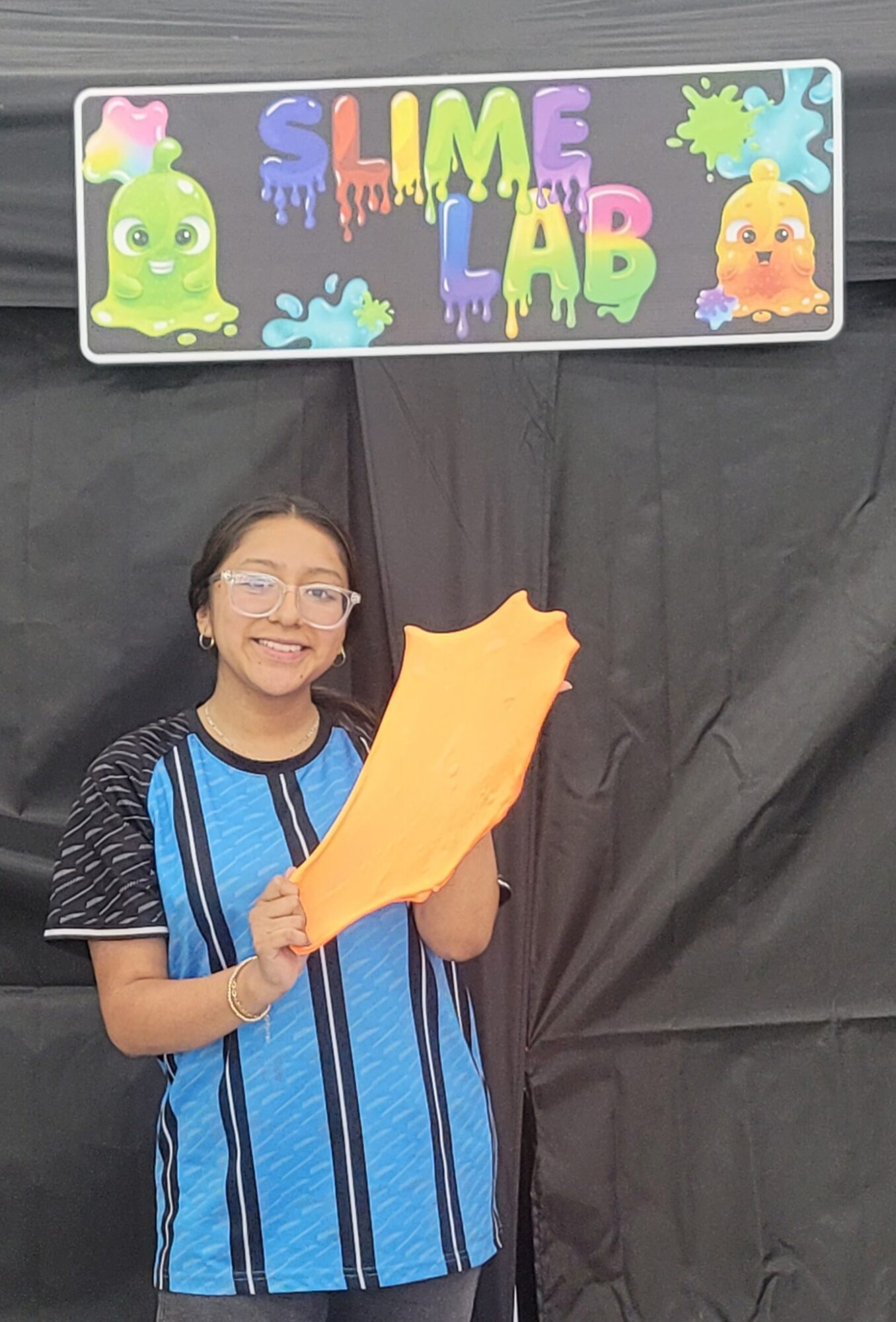 Girl holding a large orange foam object, smiling, standing in front of a black backdrop with a colorful slime lab sign.