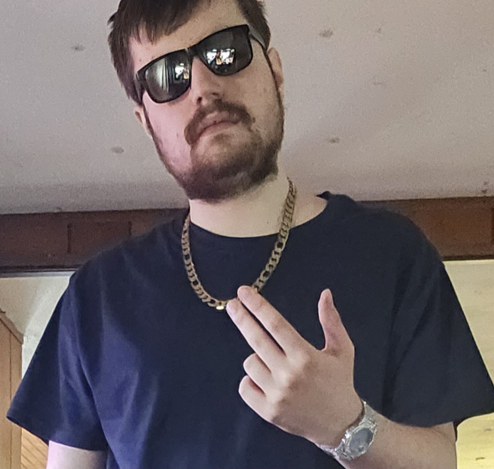 Man with sunglasses, beard, black T-shirt, gold chain, and watch making a peace sign gesture.