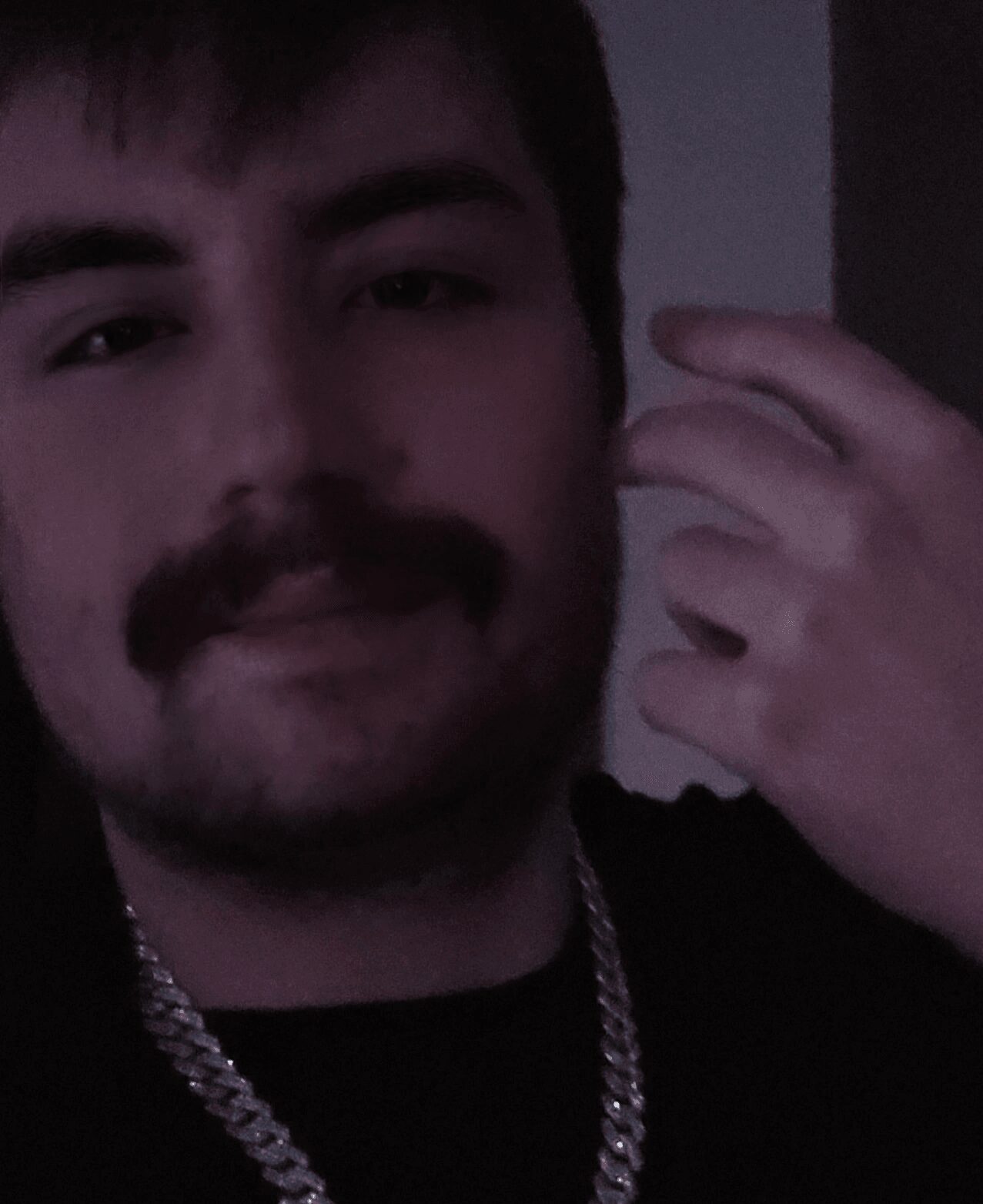 Close-up of a person with dark hair, mustache, and beard, holding their hand near their face, in a dimly lit environment.