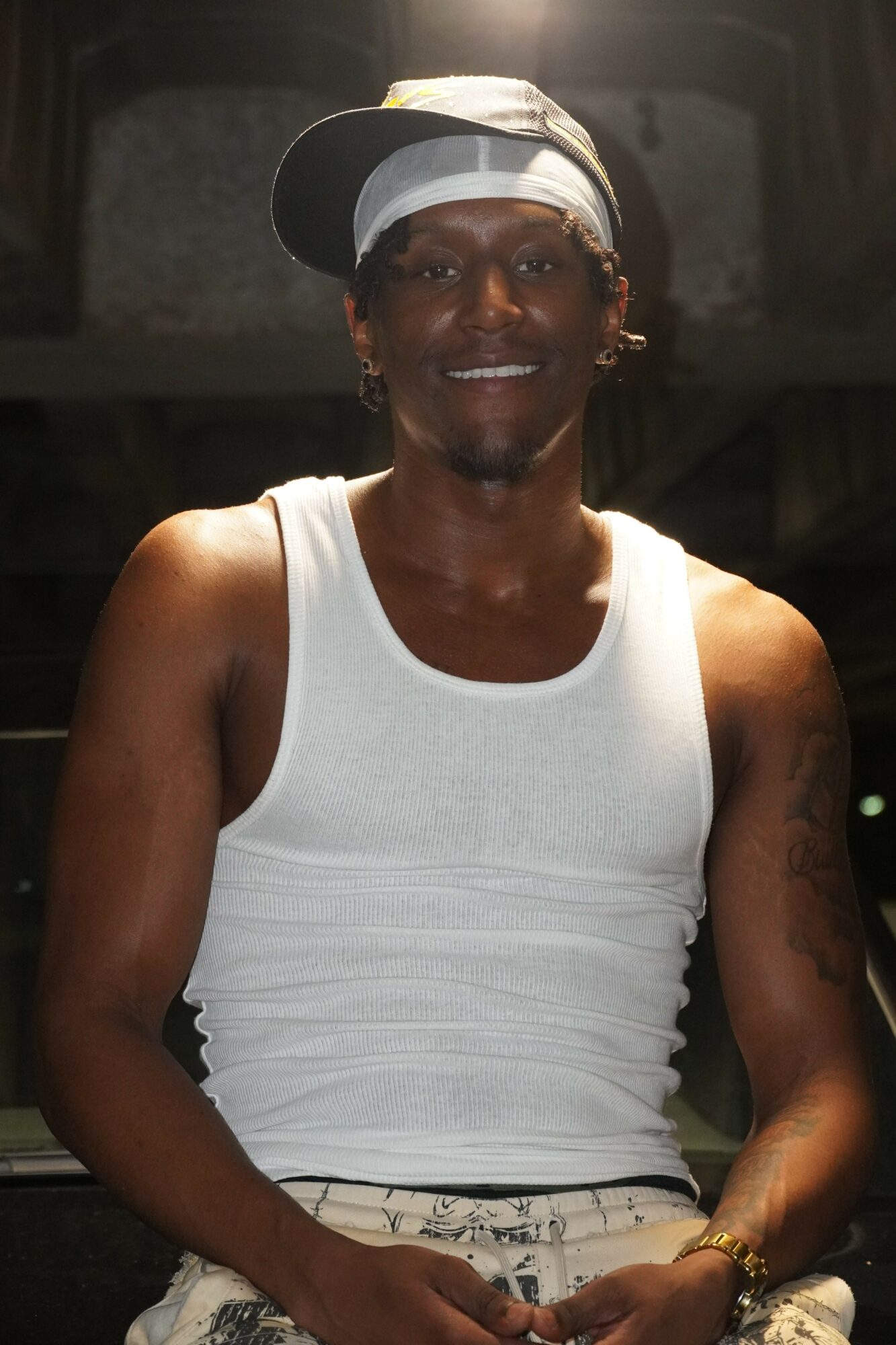 Man wearing a white tank top and a cap, smiling, in a dimly lit environment.
