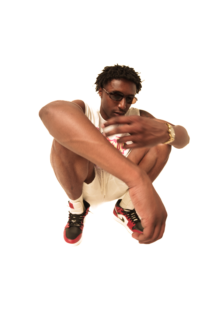 Young man with glasses crouching, arms crossed, wearing a white shirt, sneakers, and a gold watch, against a white background.