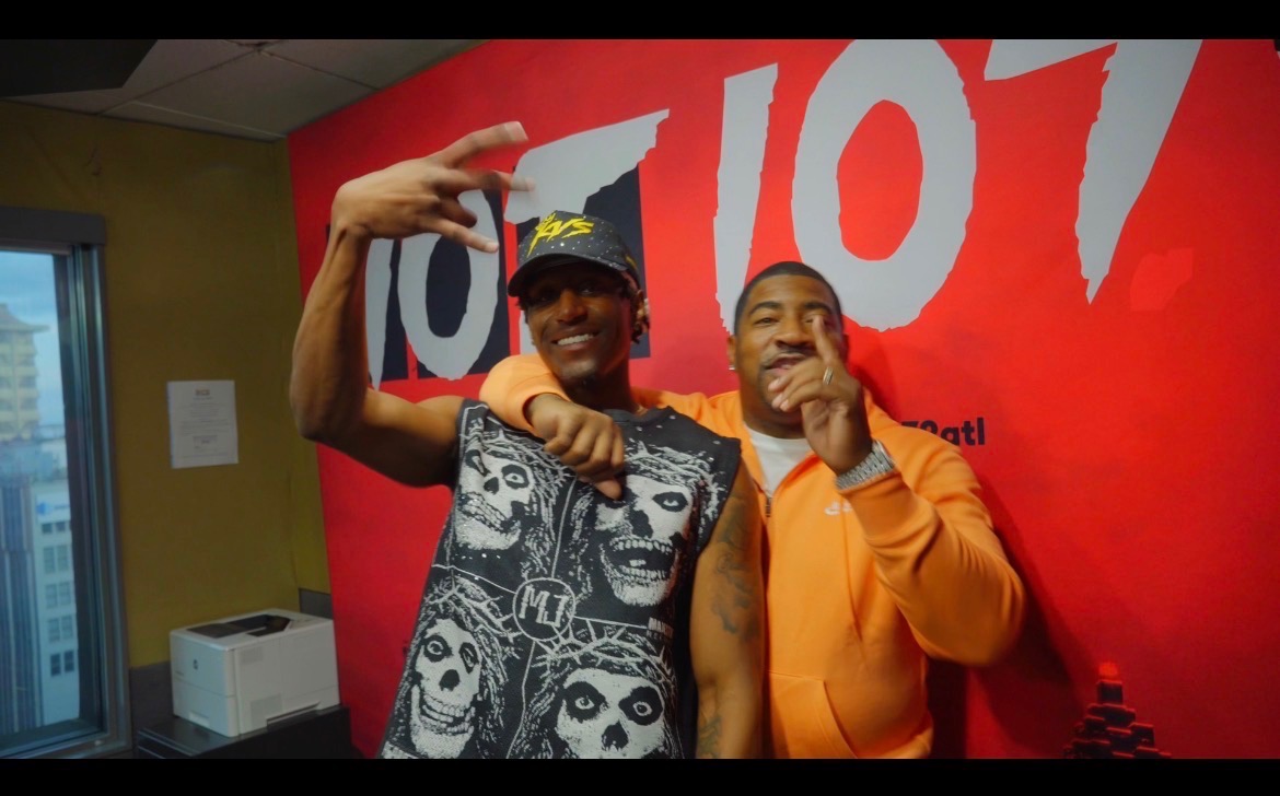 Two men smiling and posing in front of a red wall with large white text, one wearing a cap and sleeveless shirt, the other in an orange hoodie.