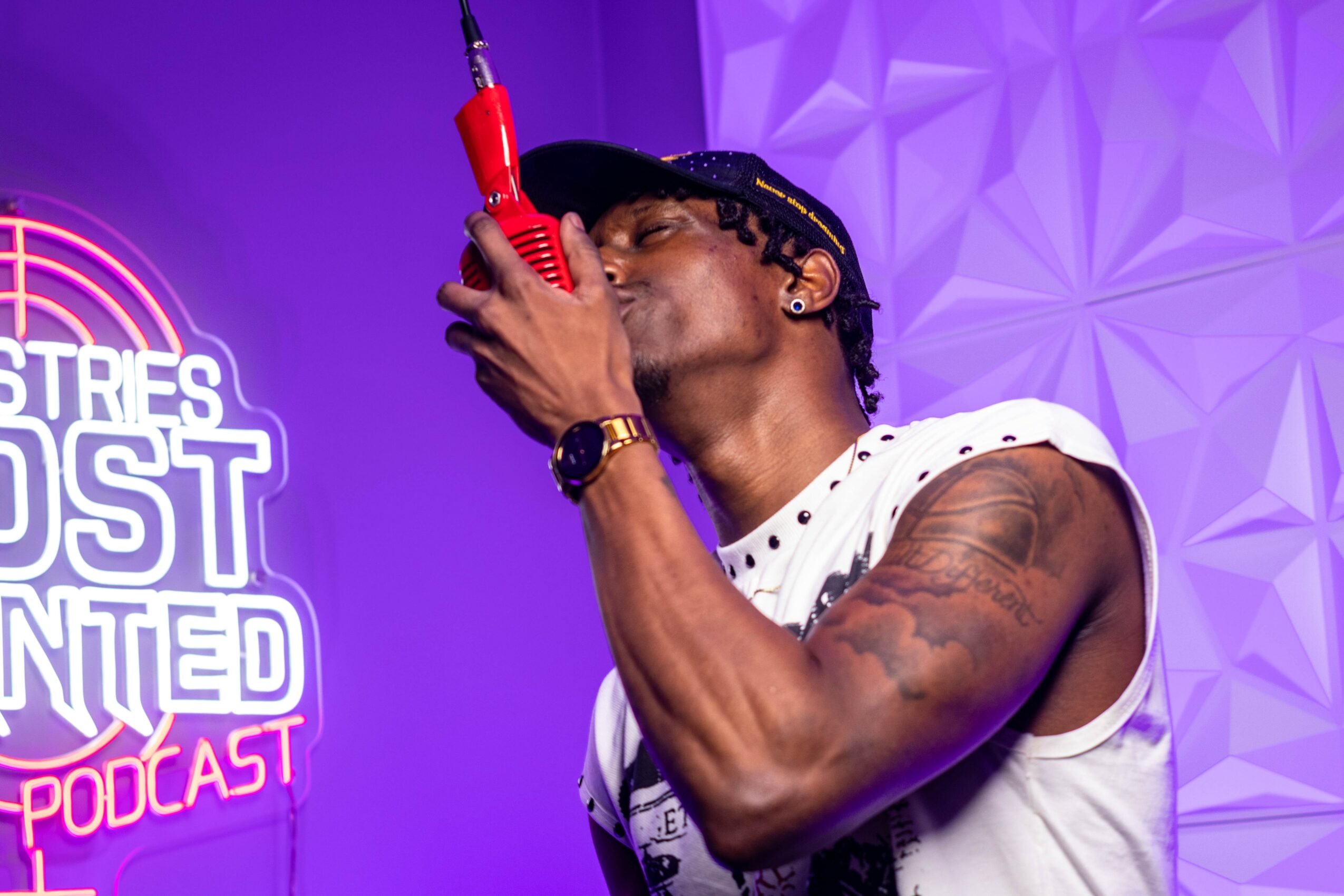 Person holding a red microphone close to face, with purple background and geometric wall pattern.