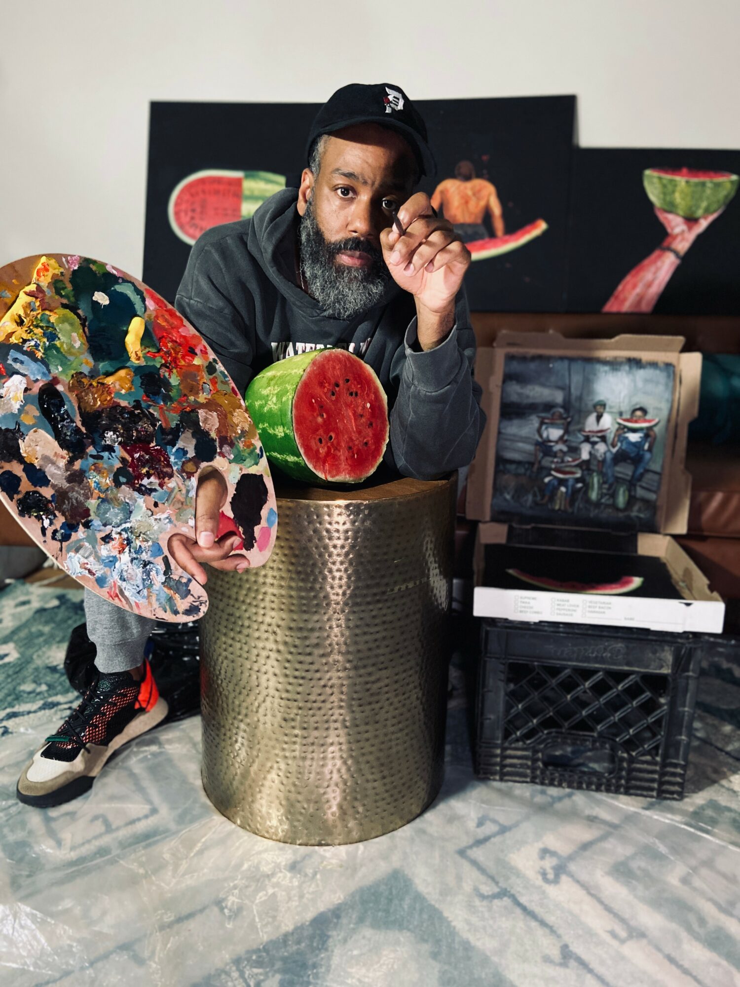 Man with a beard and cap holding a paint palette, surrounded by colorful objects, in an indoor setting.
