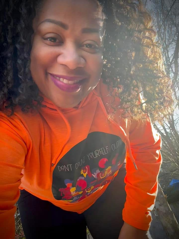 Smiling woman with curly hair wearing an orange hoodie outdoors, sunlight and trees in background.