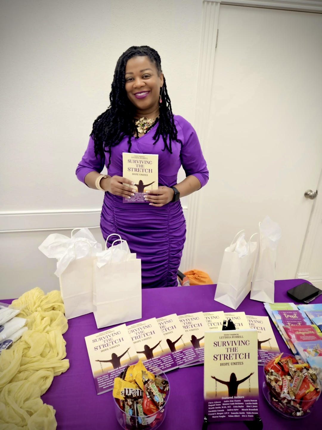 Woman in purple dress holding a book, standing behind a table with books and gift bags, smiling indoors.