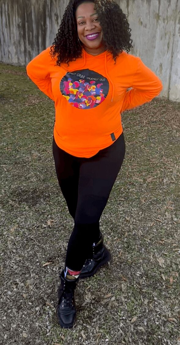 Woman standing outdoors on grass, wearing an orange sweatshirt with a colorful design, black pants, and black shoes.