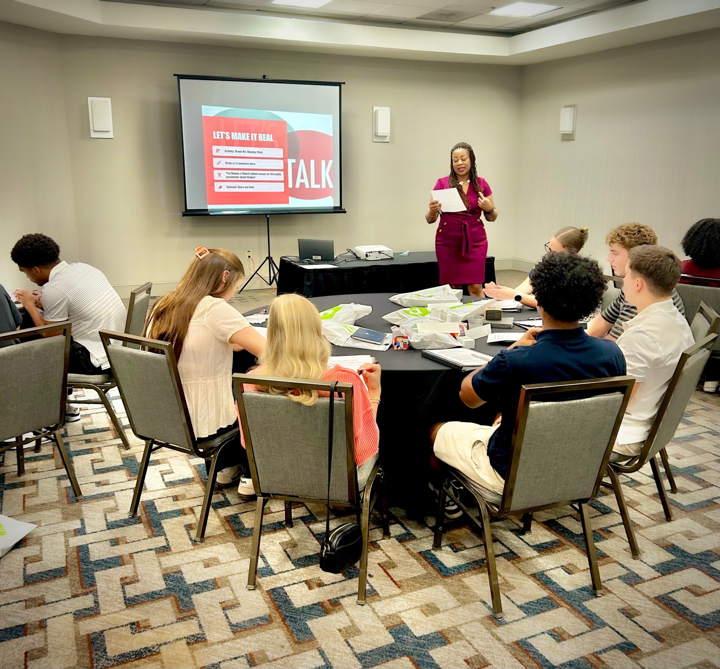 Woman standing and speaking to a group of children seated at a round table in a conference room, with a presentation screen behind her.
