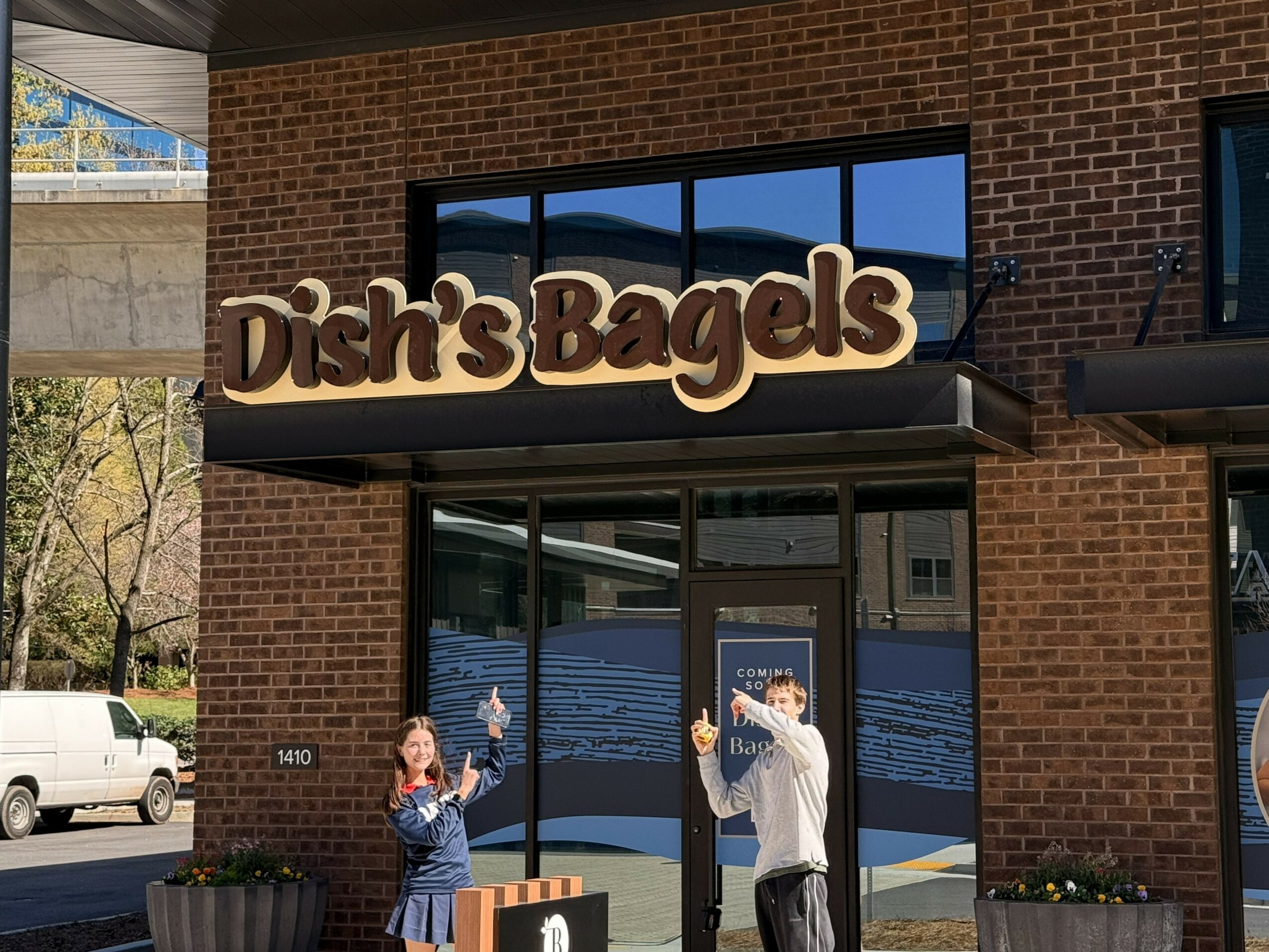 Dish's Bagels storefront with a sign, brick exterior, and large windows, two people waving outside, and cars parked nearby.