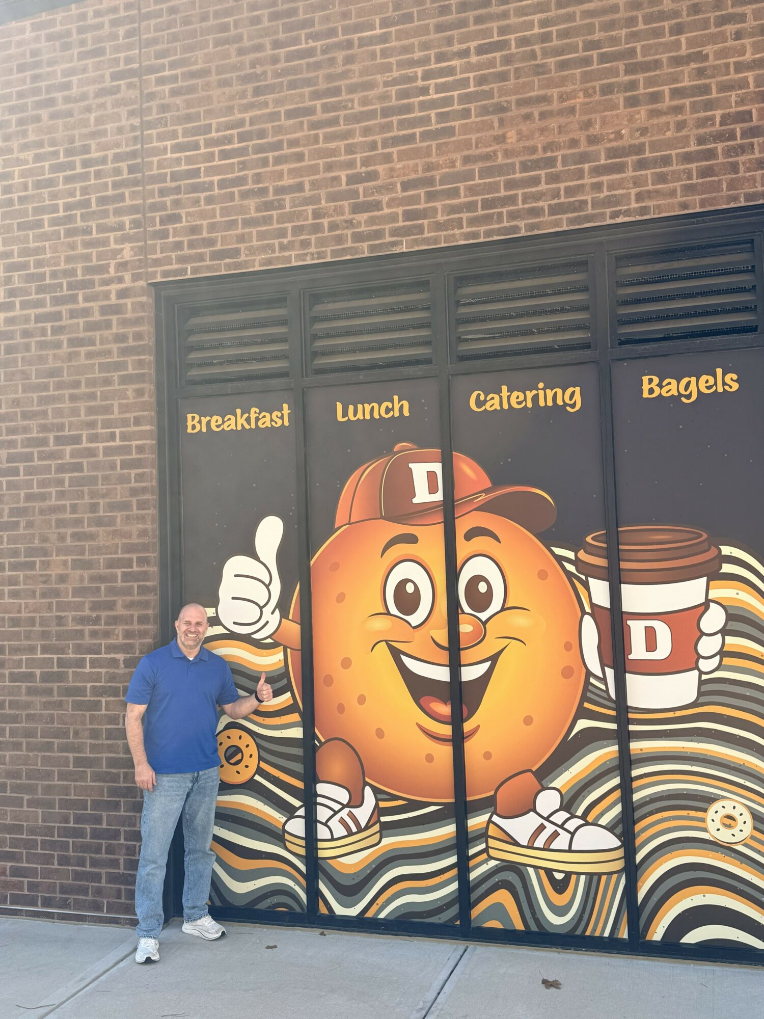 Person standing next to a colorful cartoon bakery window display with a smiling baked good character, coffee cups, and text.