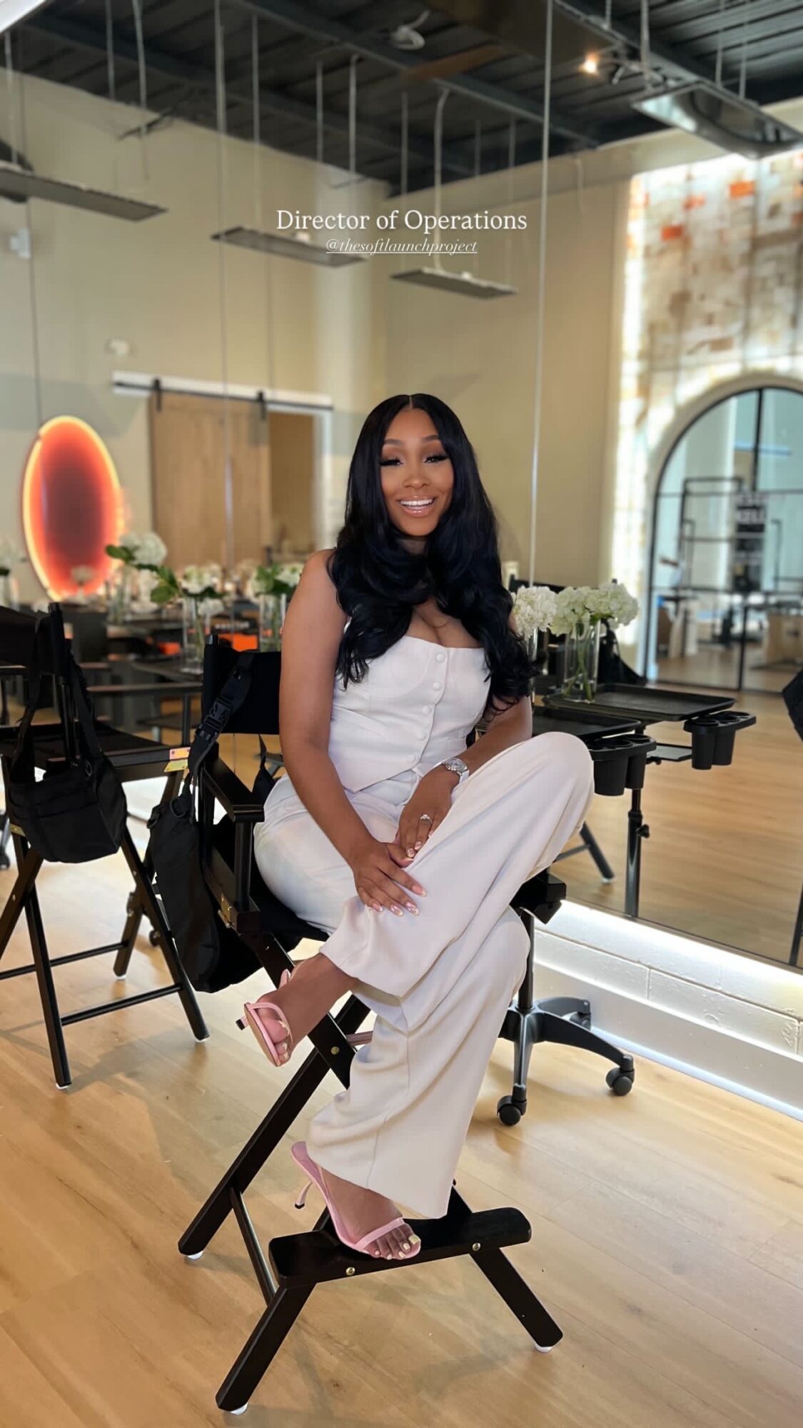 Woman sitting on a black chair in a room with wooden floors, flowers, and large windows, smiling at the camera.
